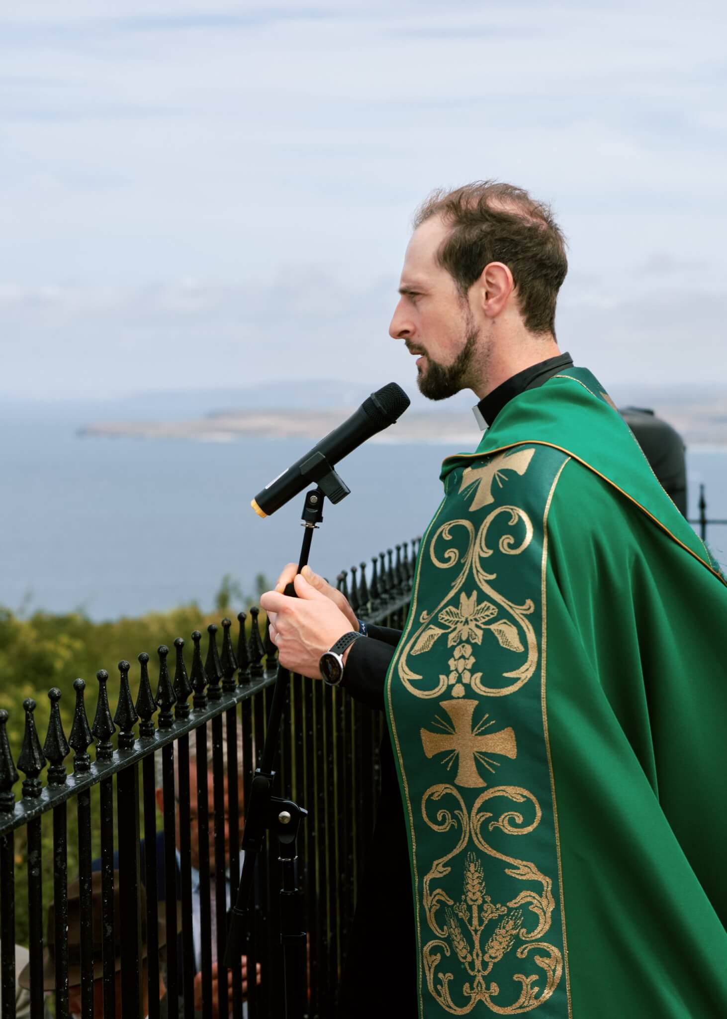 The Vicar of St Ives in green vestments speaks into a microphone with the sea behind