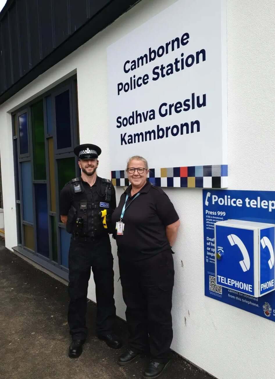 A police officer and a PCSO stand smiling outside Camborne Police Station beneath the bilingual English and Cornish sign reading Sodhva Greslu Kammbronn