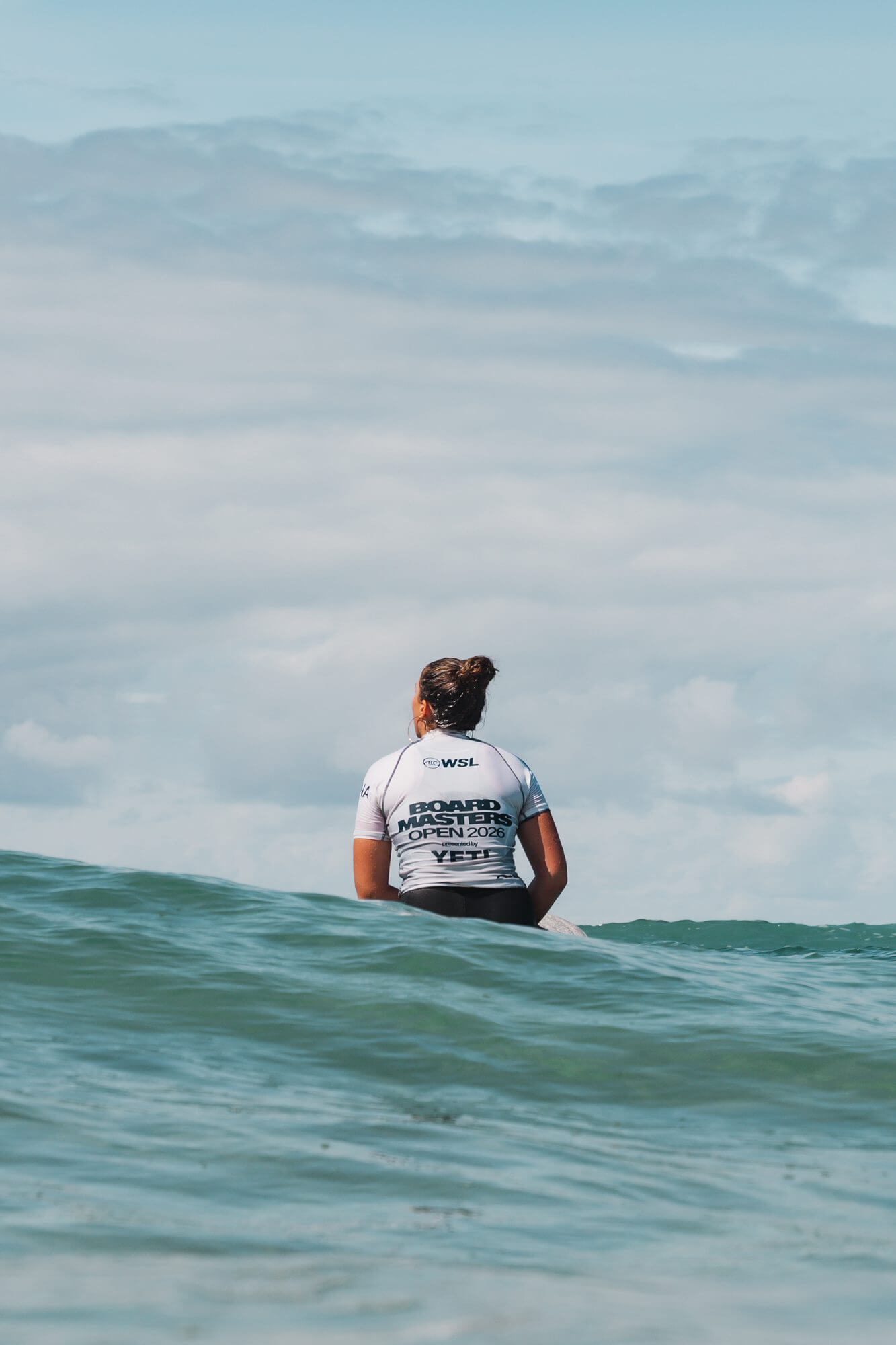 A surfer in a Boardmasters Open 2026 jersey sits on her board waiting for a wave