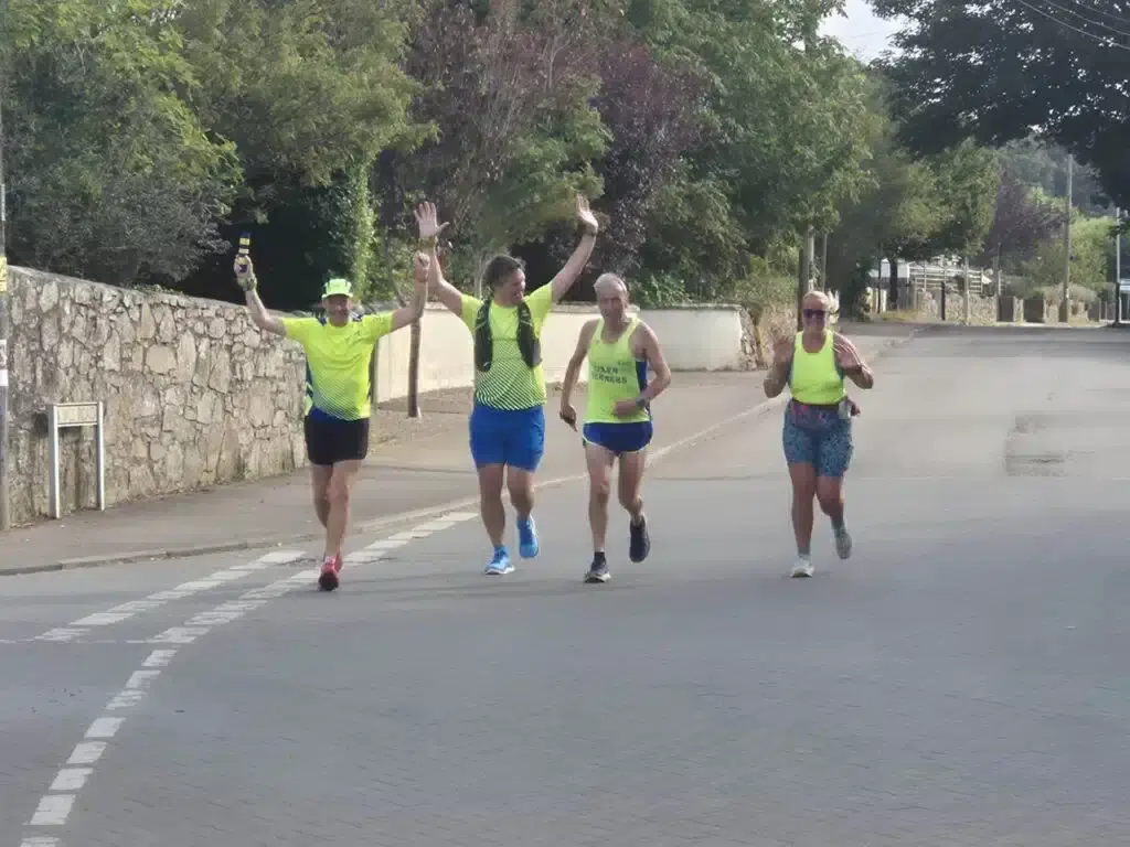 Four Carn Runners cheering with arms aloft as they run along a road