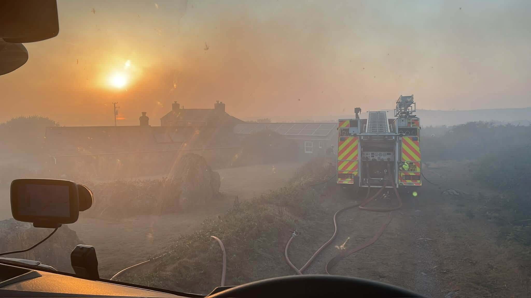 A fire engine and hoses at a smoky rural scene as the sun sits low on the horizon