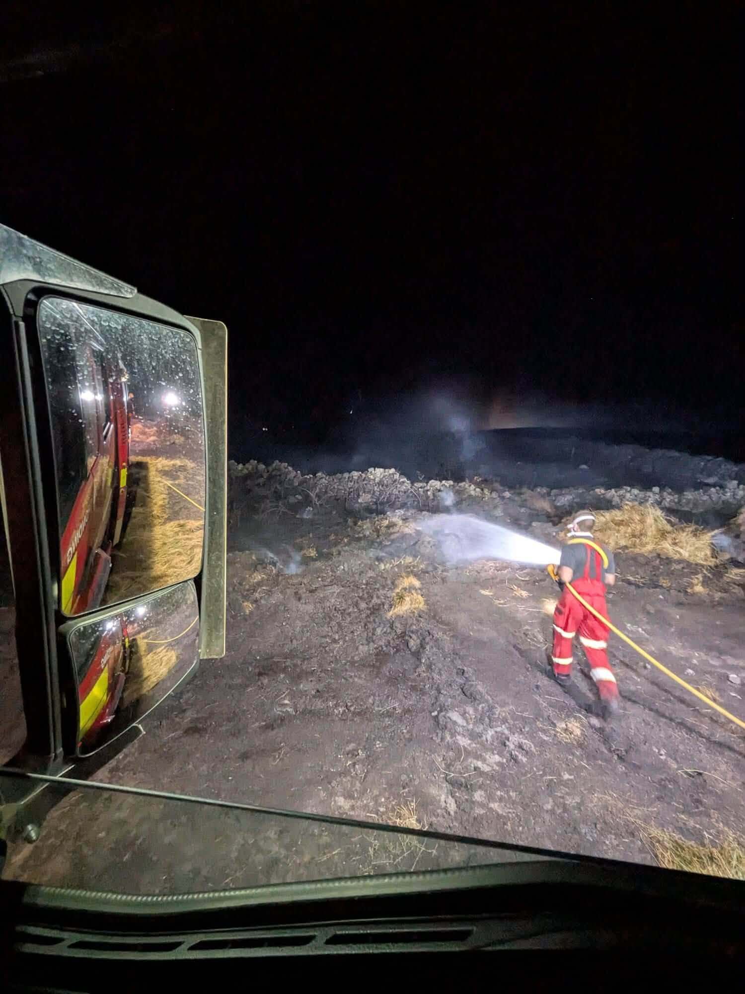 A firefighter hosing down scorched ground in the dark beside a fire engine