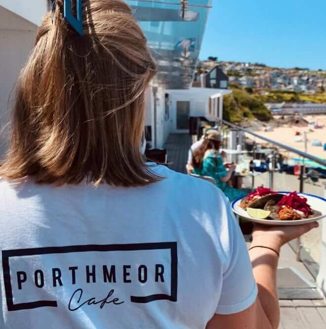 A cafe worker carrying a plate of food on a terrace above a St Ives beach