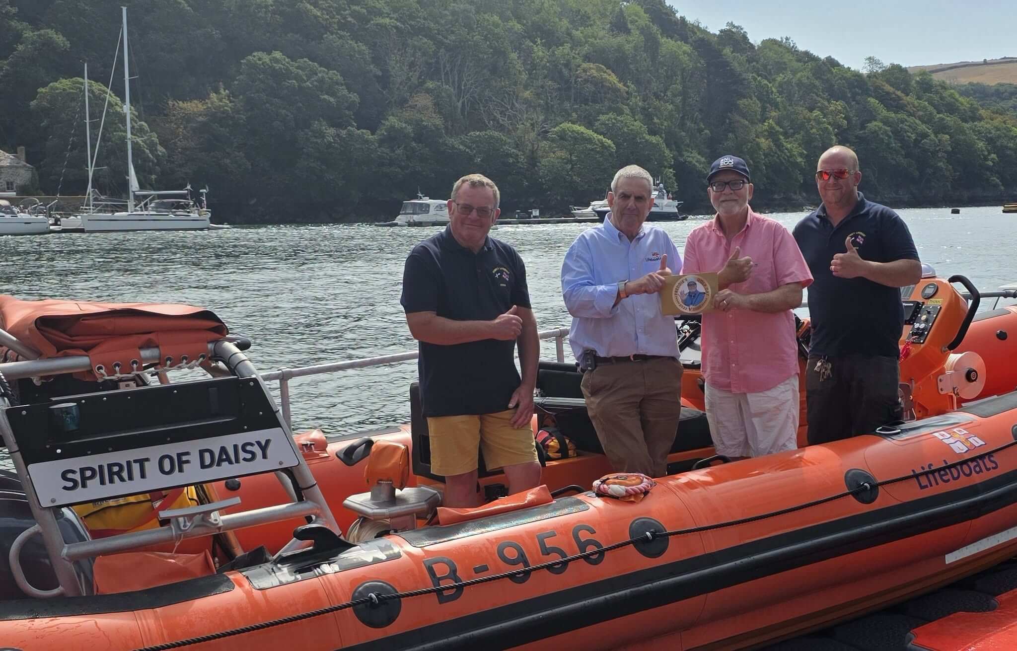 Four men standing beside Fowey RNLI's orange inshore lifeboat on the water, giving thumbs up as a cheque presentation plaque is held up