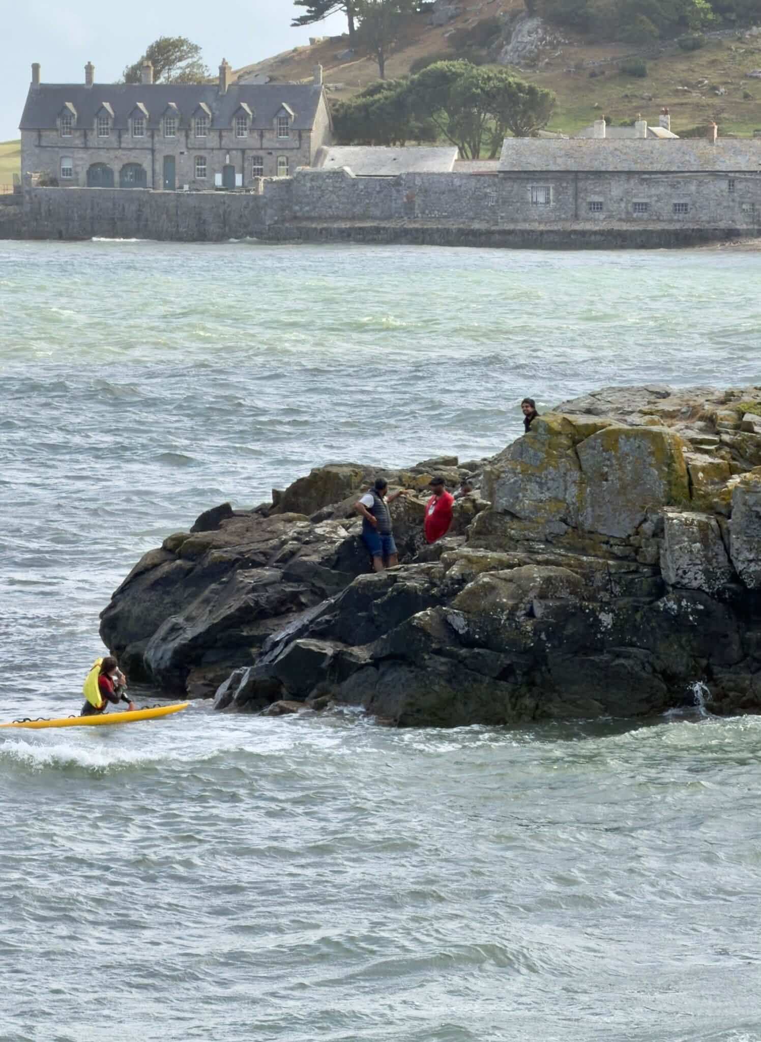 People wait on rocks off Marazion as an RNLI lifeguard approaches on a rescue board, with St Michael's Mount buildings in the background