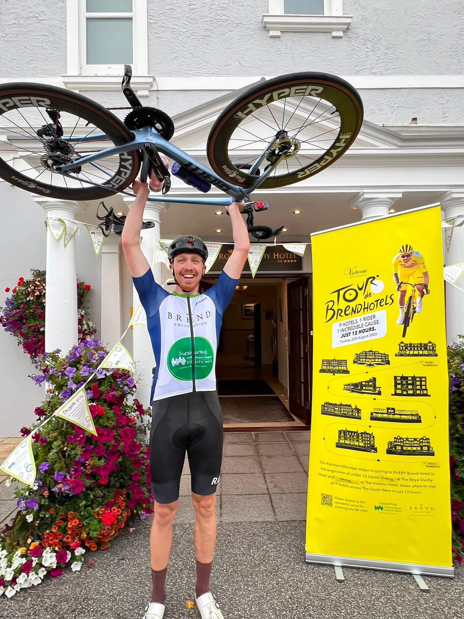 Aidan holding his bike aloft outside The Royal Duchy Hotel in Falmouth, with the Tour de Brend Hotels banner alongside