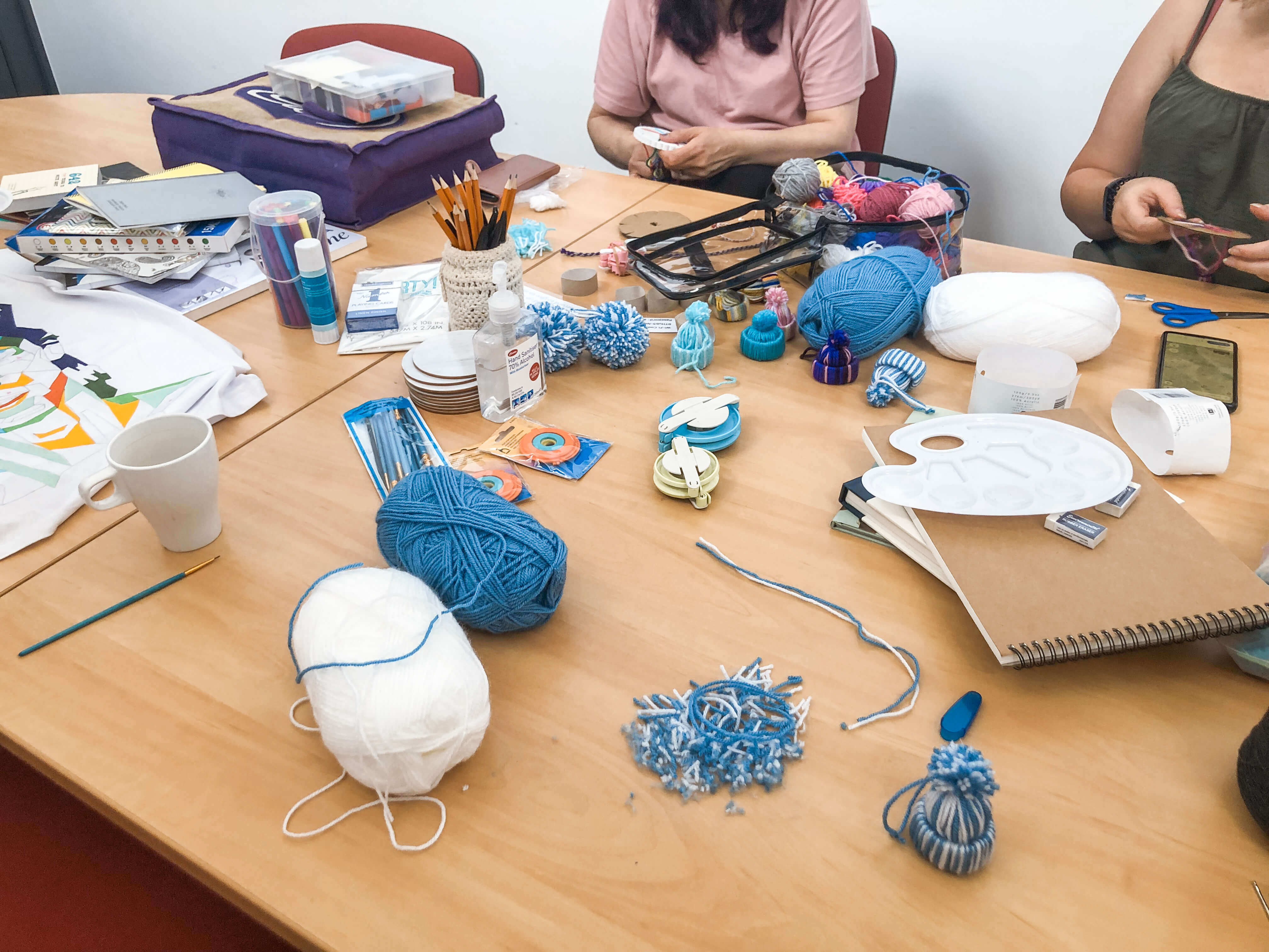 Hands stitching a cross stitch square at a table covered with patterns, thread and coloured pencils