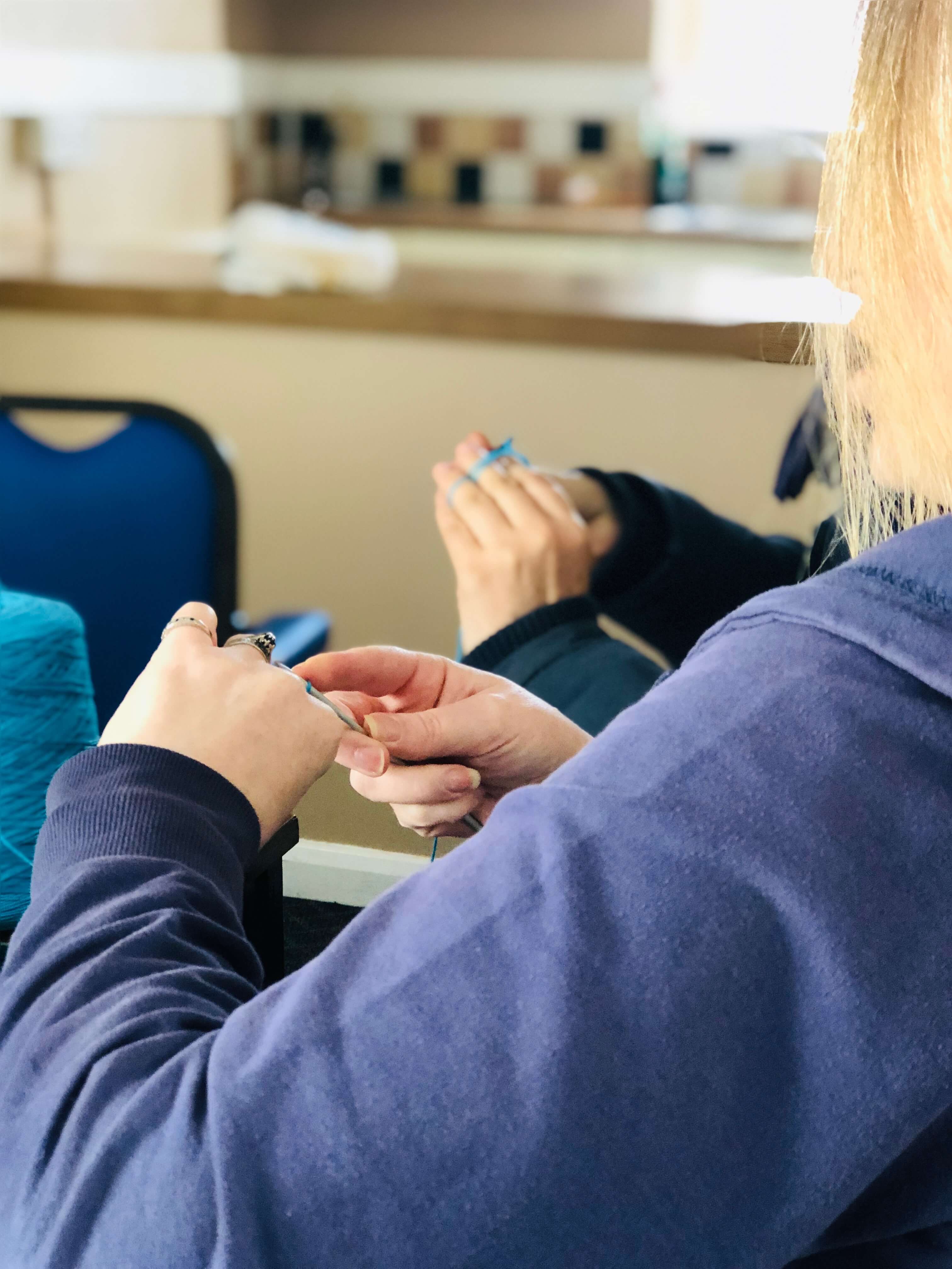 Two people crocheting with blue yarn, seen over the shoulder of one of them