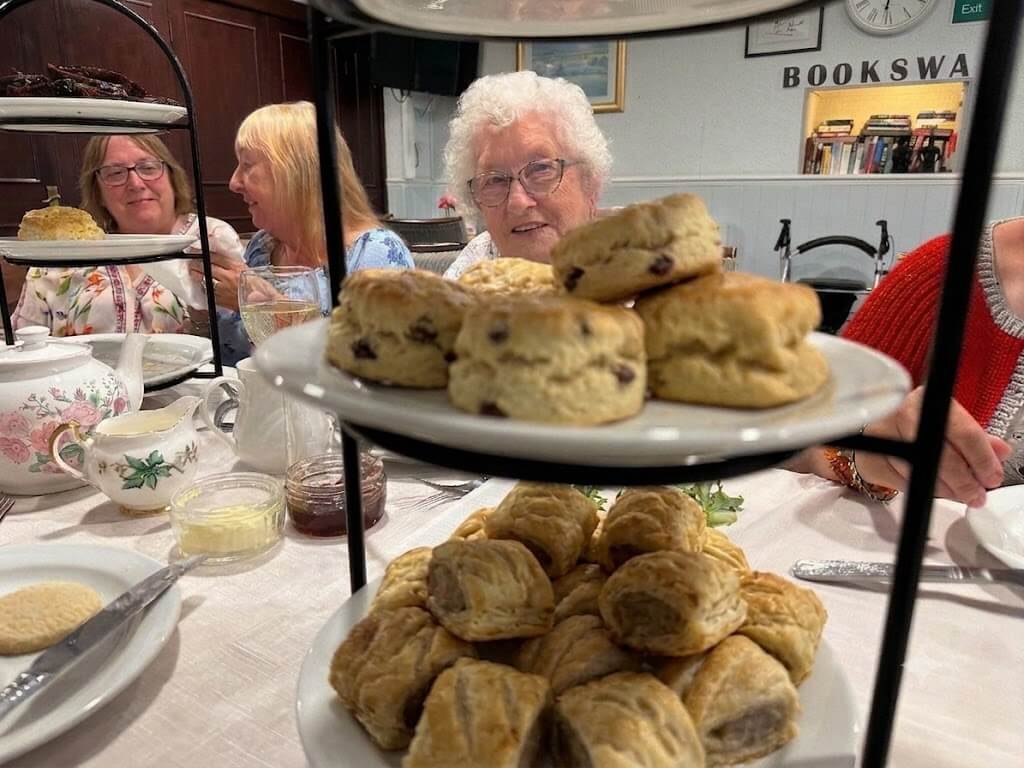 A tiered stand of scones and sausage rolls at the Oddfellows afternoon tea, with a member smiling behind
