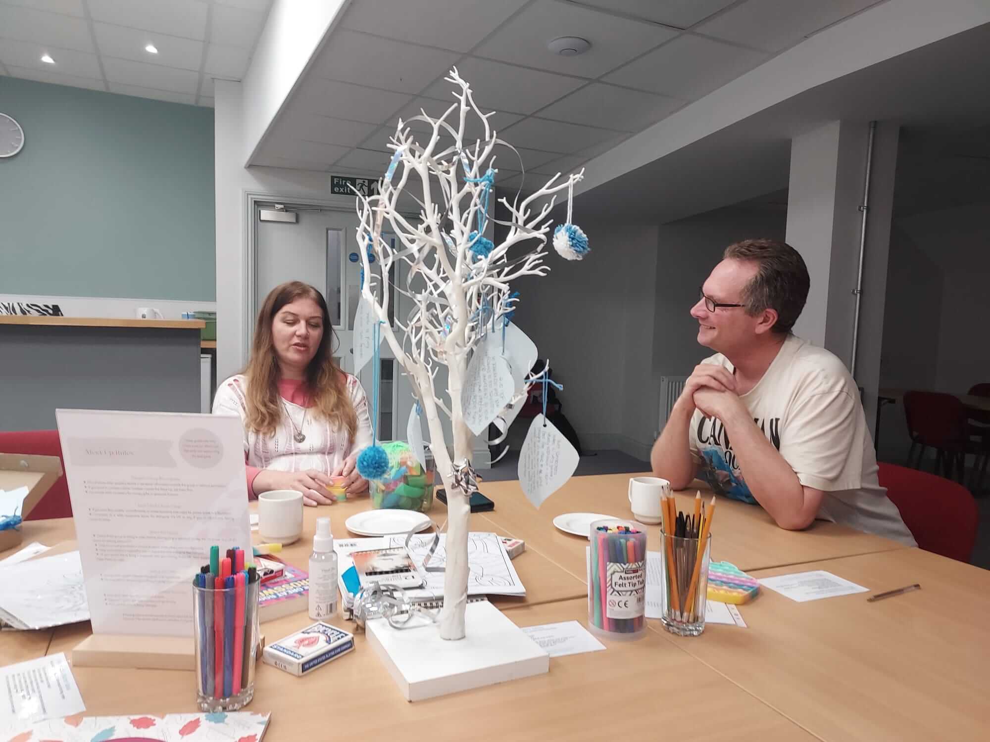 Two people sitting either side of a craft table with a white wish tree at a Quiet Connections Meet Up