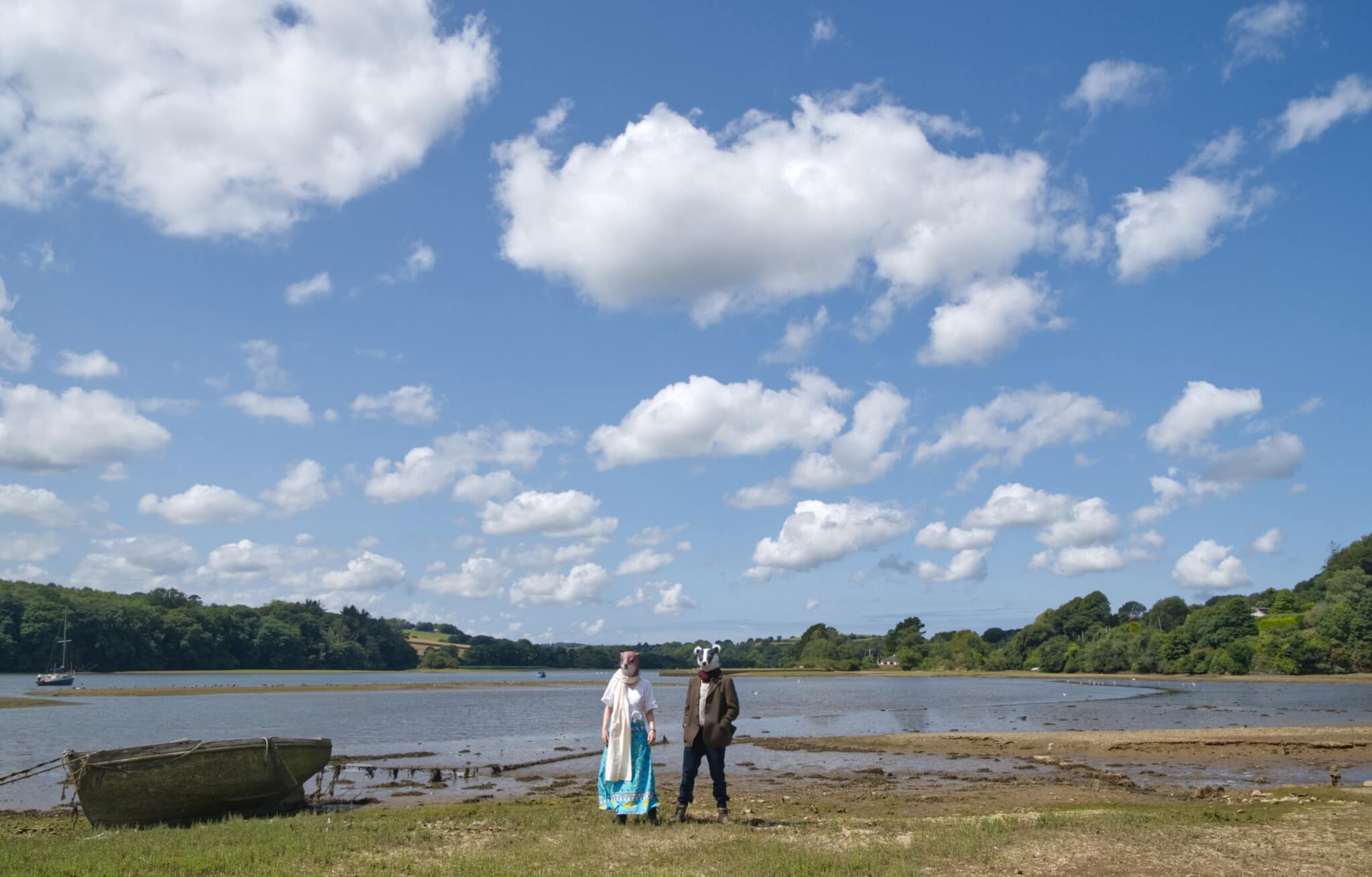 Chris and Annabelle South of Meles Lutra in otter and badger masks beside a wooden boat on a Cornish estuary at low tide