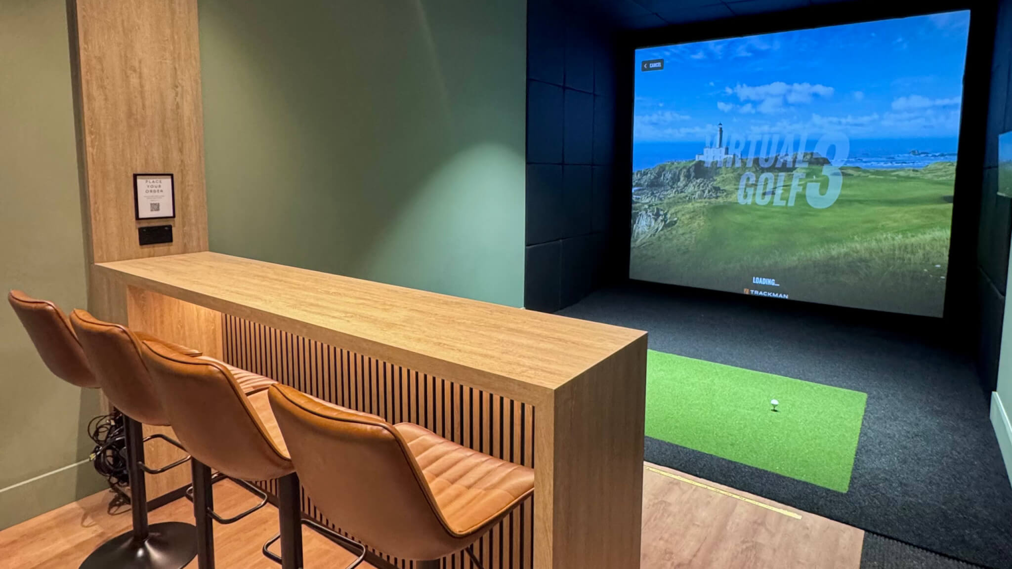 The new TrackMan golf simulator studio at Carlyon Bay Golf Club, with bar seating, a screen showing a coastal course and a putting mat