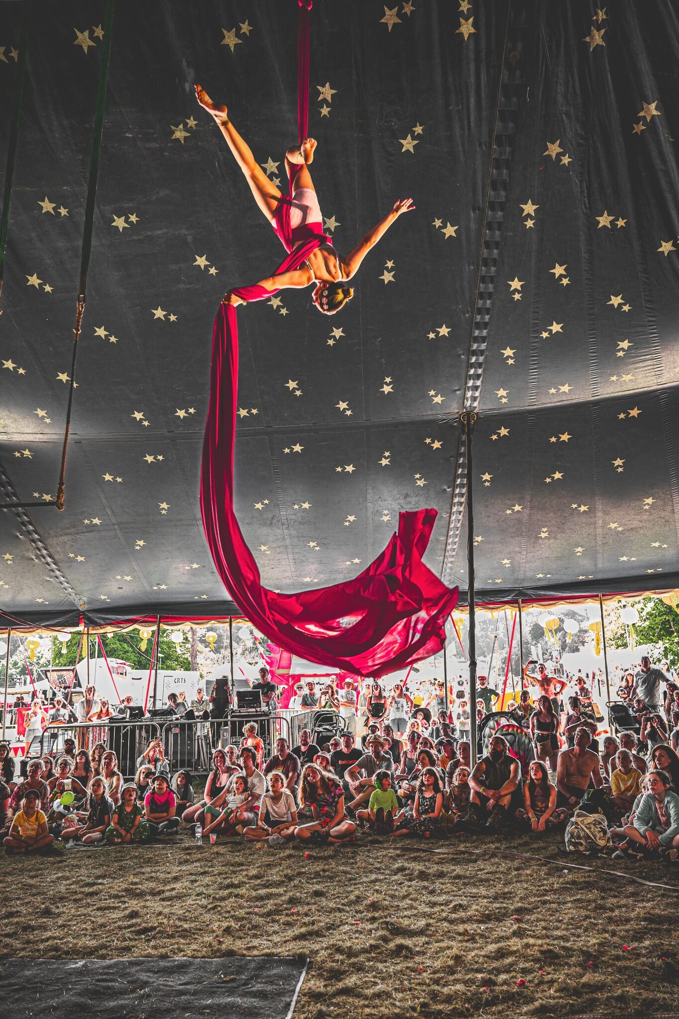 Aerial silks performer hanging upside down in a star-covered big top above a seated crowd