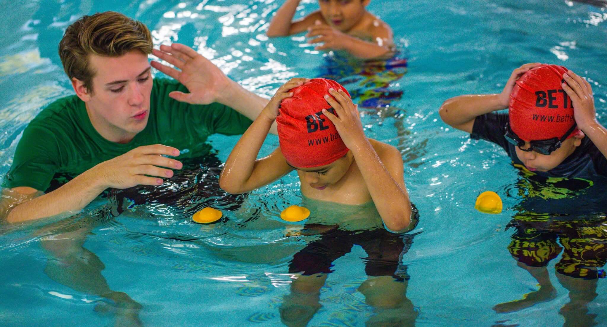 A swimming teacher instructing young children wearing red Better swim caps in a leisure centre pool