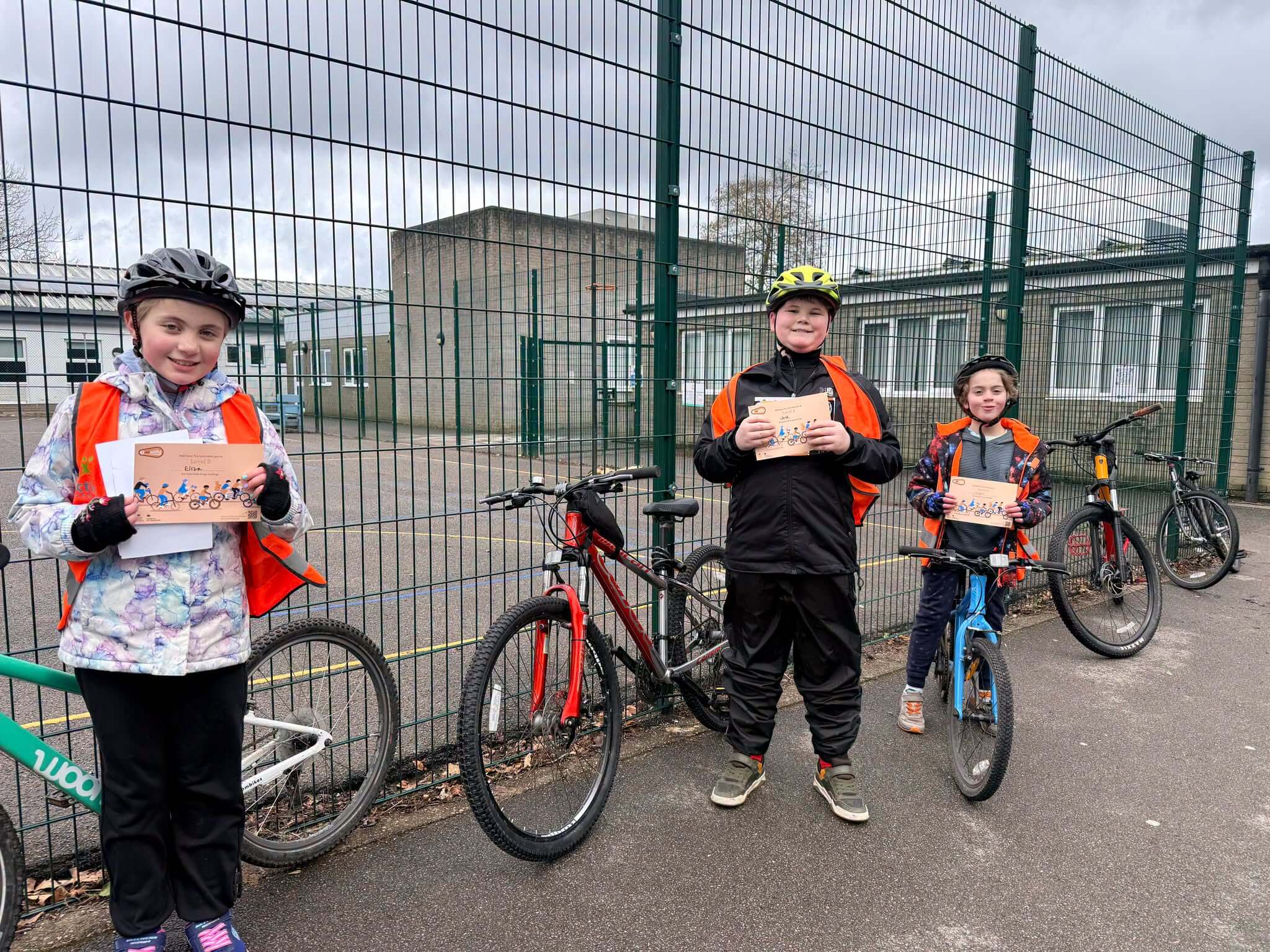 Three young cyclists in hi-vis holding Bikeability certificates beside their bikes