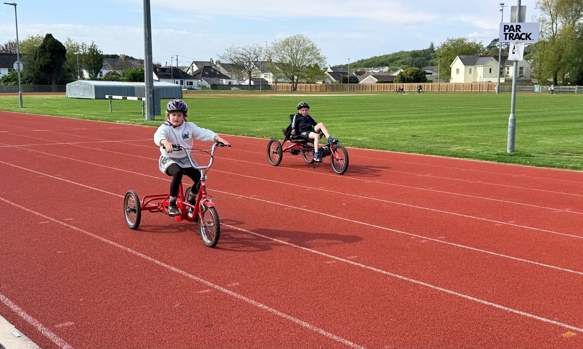 Two children riding a tricycle and an adapted recumbent cycle on the red running track at Par Track