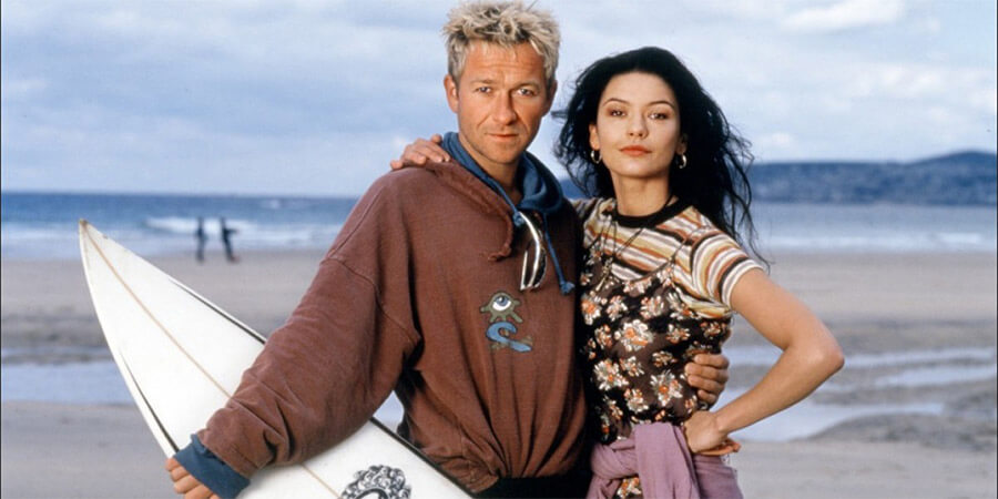 Two characters from the film Blue Juice on a Cornish beach with a surfboard