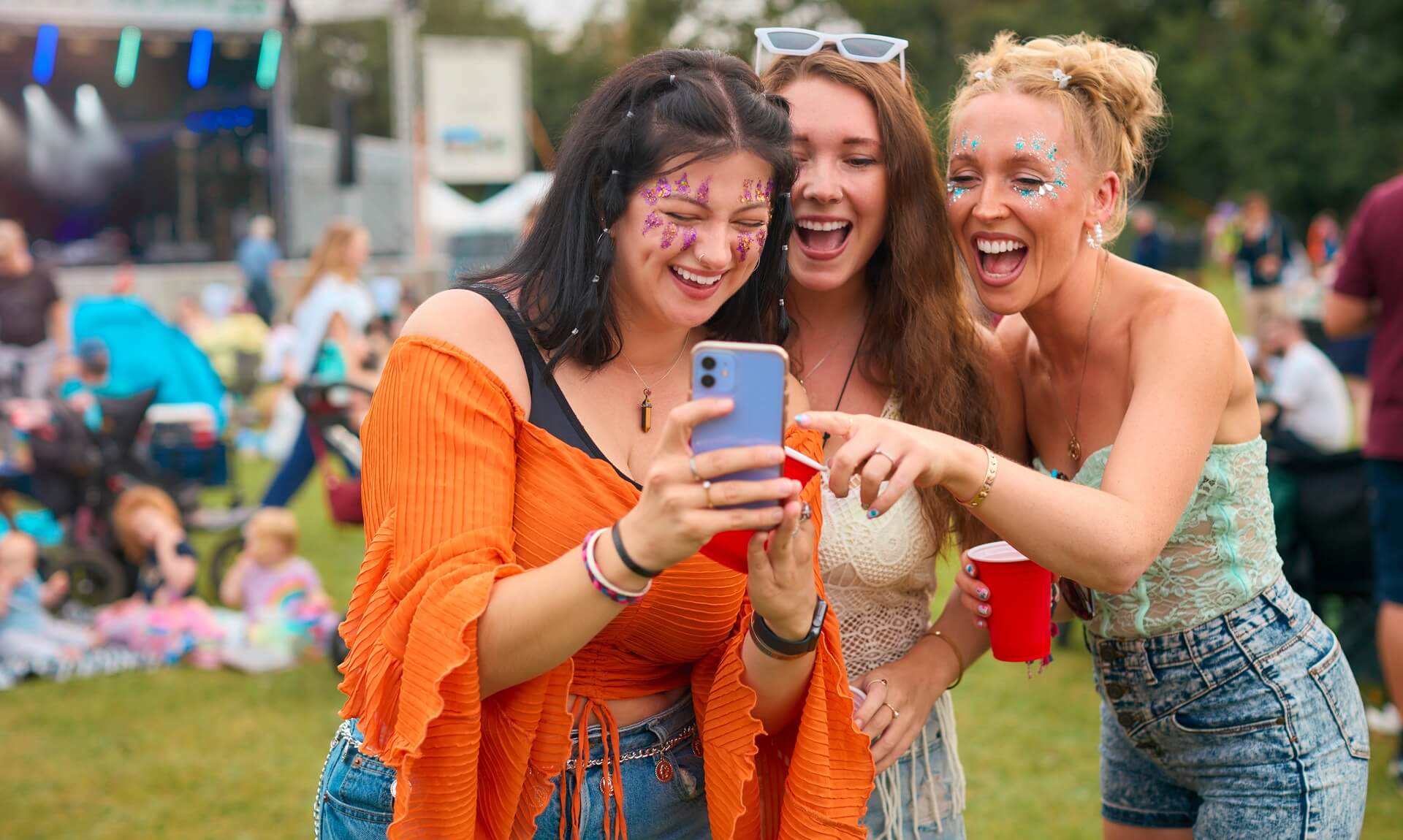 Three friends with festival glitter laughing at a phone screen at Boardmasters