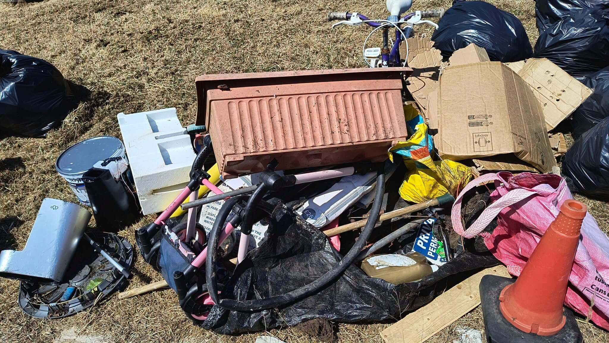 A pile of fly-tipped junk including a terracotta planter, a child's bike, a traffic cone and cardboard on dry grass