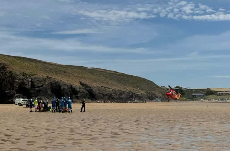 Rescue team in blue on Porthcothan beach with a red air ambulance helicopter and cliffs behind