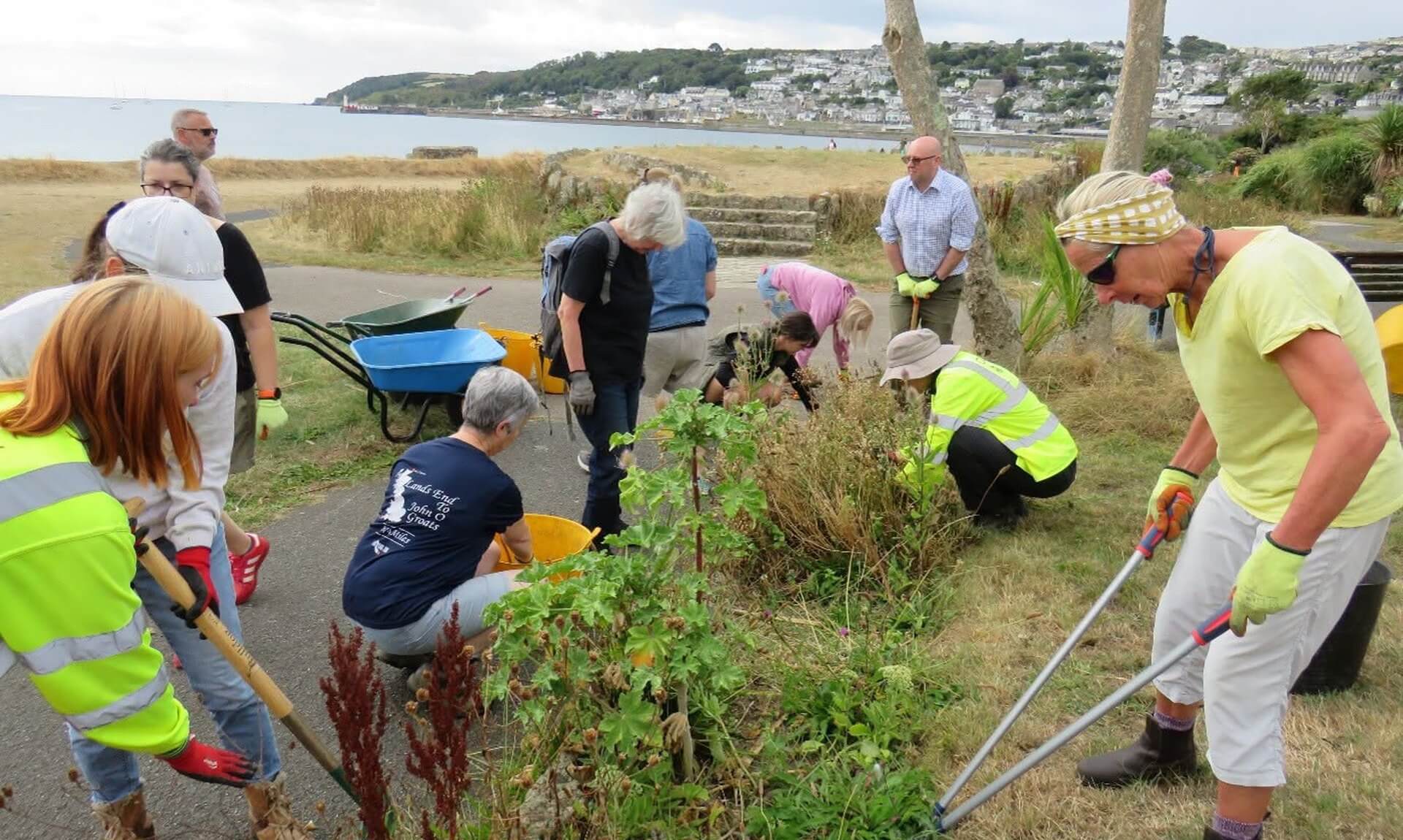 Volunteers weeding a seafront flower bed at Bolitho Gardens with Penzance across the bay behind