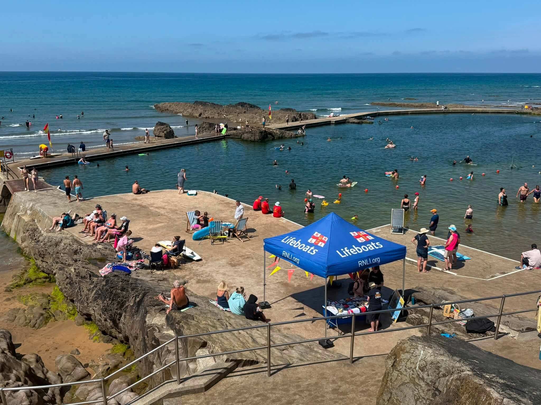 Aerial view of Bude Sea Pool packed with swimmers and the RNLI Lifeboats gazebo on the deck