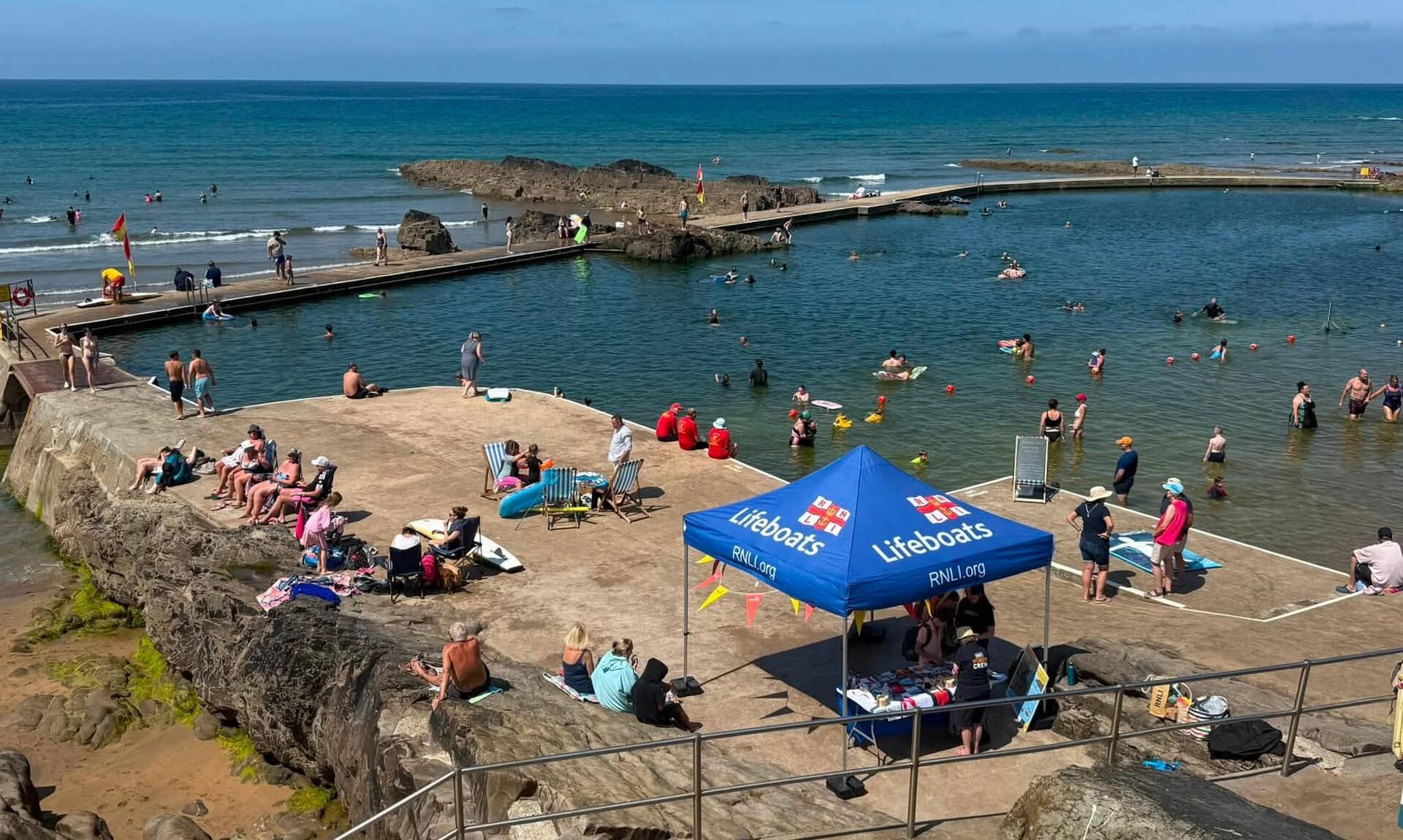 Crowds floating and gathered at Bude Sea Pool with the RNLI gazebo on the poolside