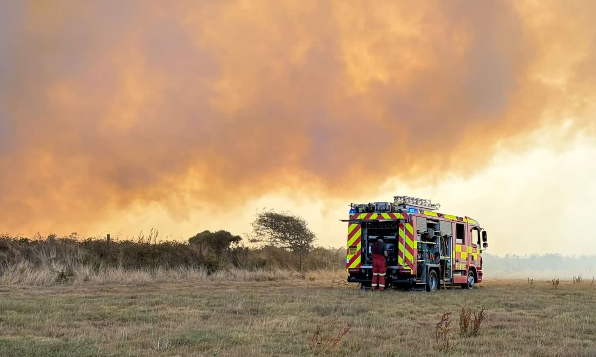 Fire engine parked in a dry field beneath a huge wall of orange wildfire smoke at Chapel Carn Brea