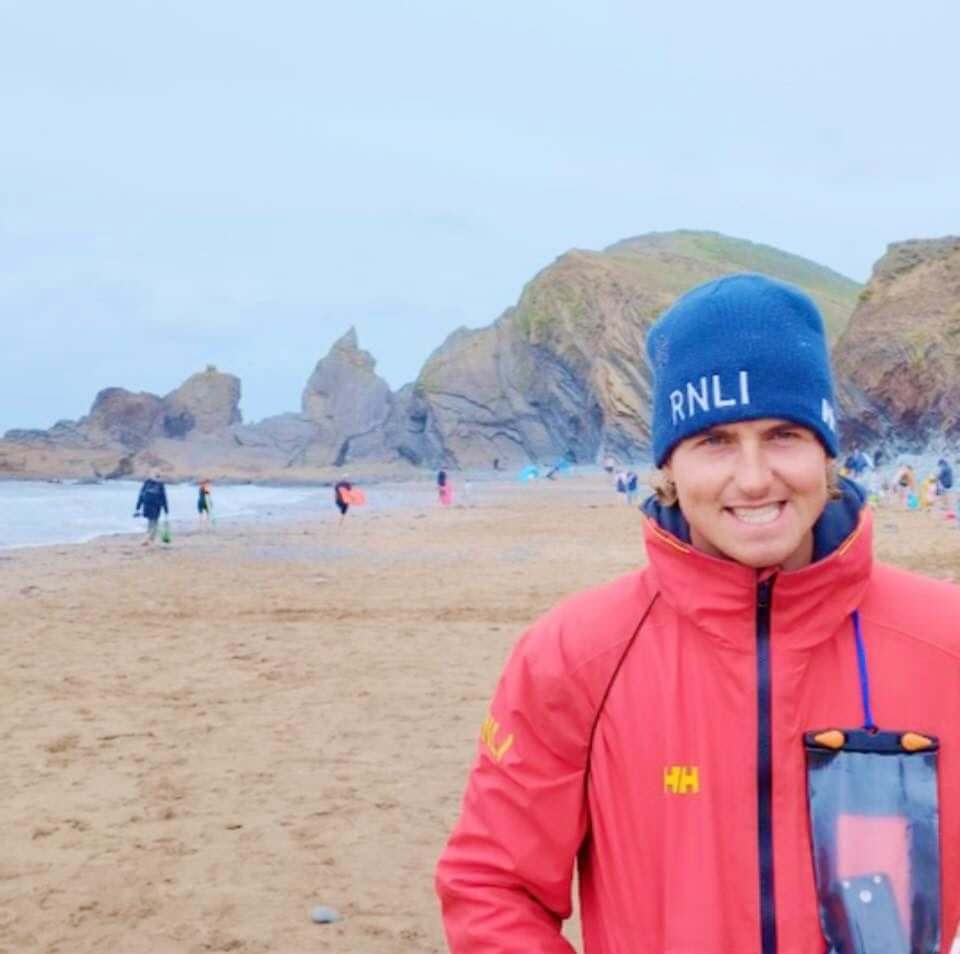 Bude senior RNLI lifeguard Charlie Barnett on the beach in his red RNLI jacket and blue RNLI beanie