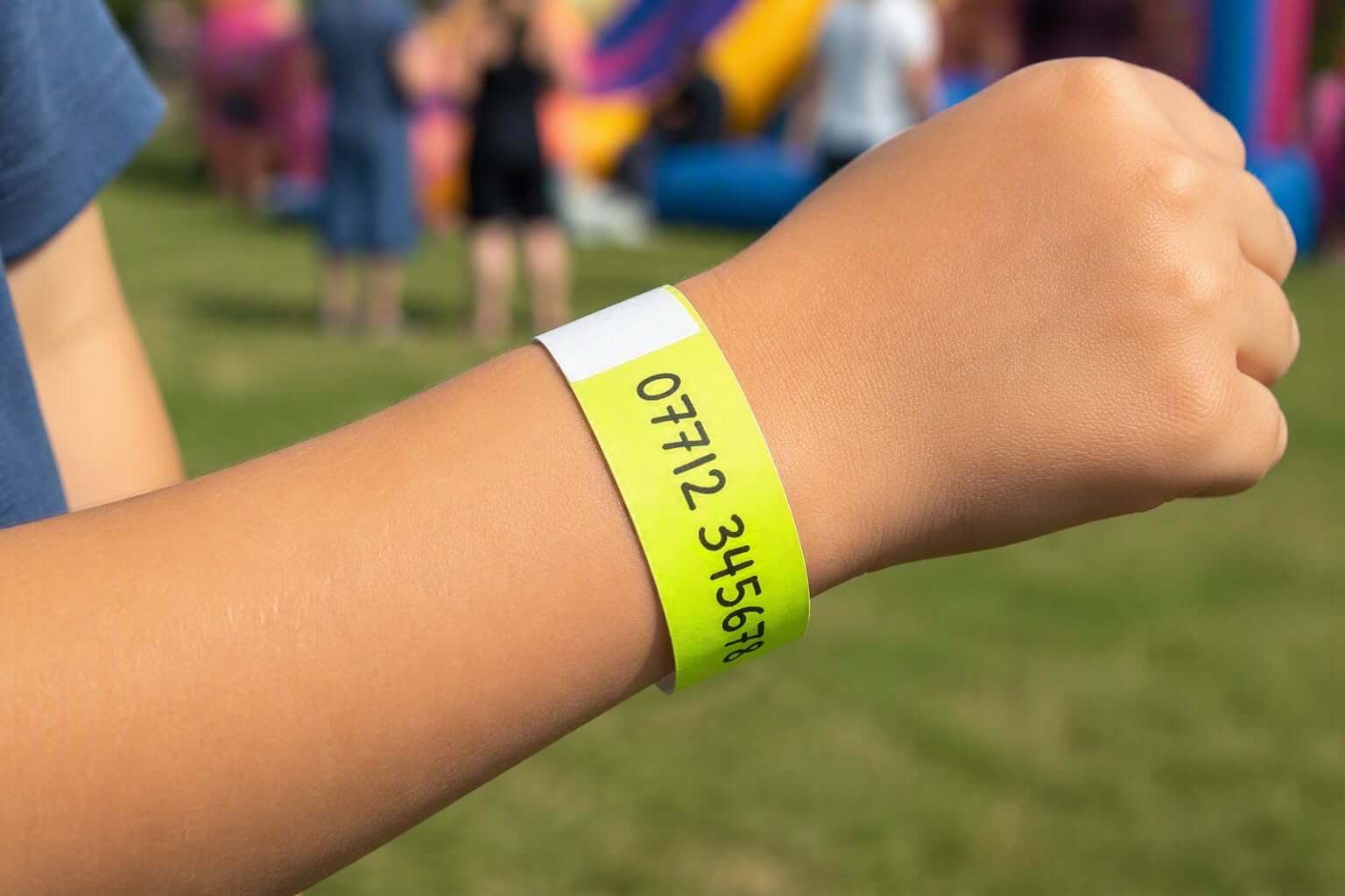A child's wrist with a yellow event wristband, a mobile phone number written on it, at a busy outdoor community event