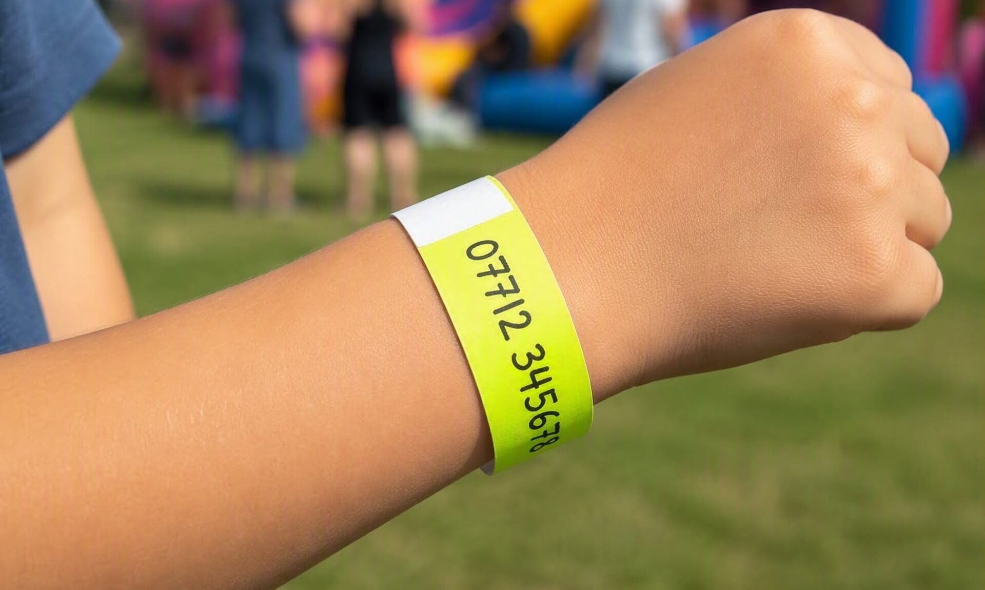 A child's wrist with a yellow event wristband, a mobile phone number written on it, at a busy outdoor community event
