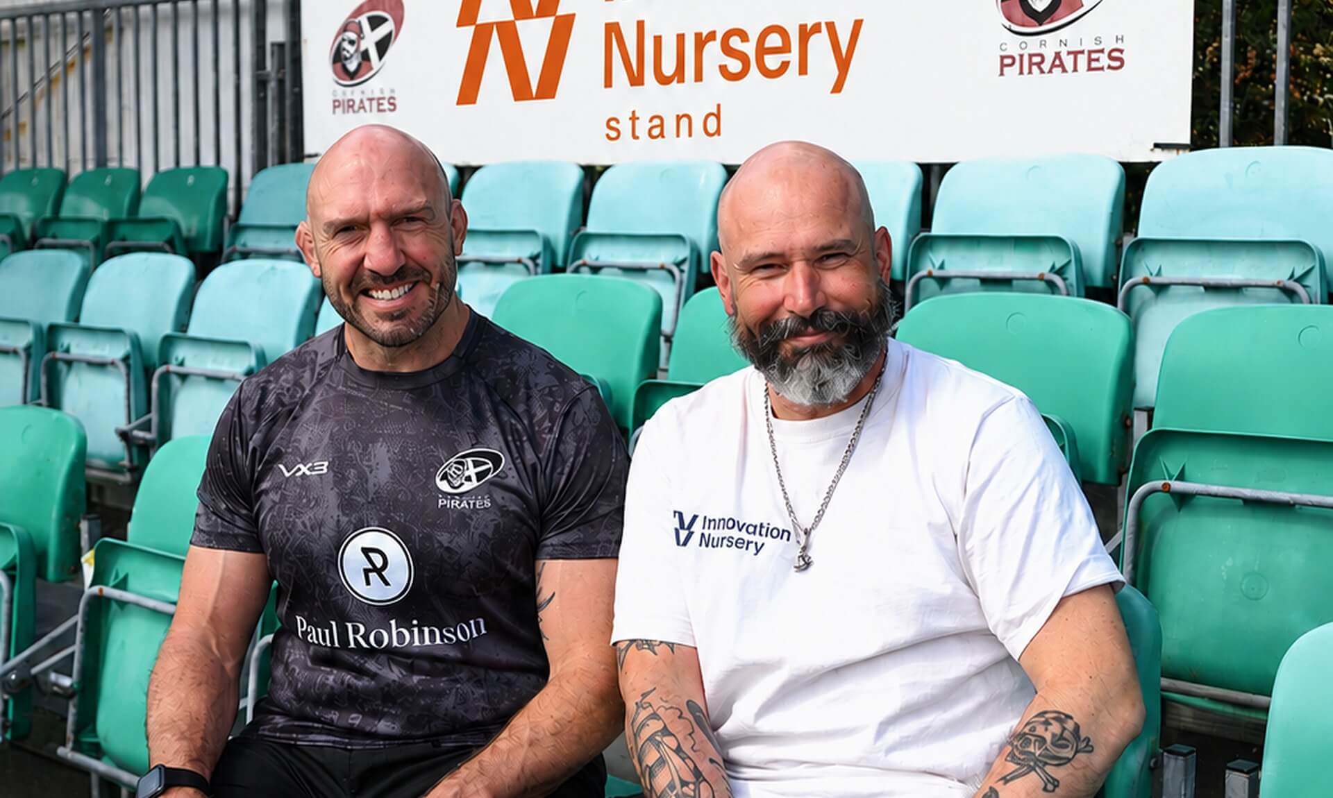 Two men sitting in stadium seating below the newly branded Innovation Nursery stand sign at the Cornish Pirates