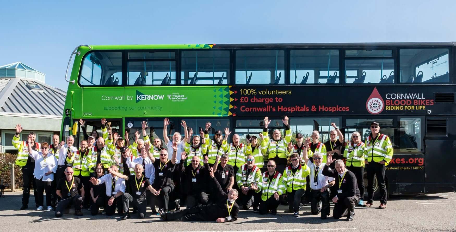 Cornwall Blood Bikes volunteers in hi-vis jackets cheering in front of a Kernow double decker bus in the charity's livery