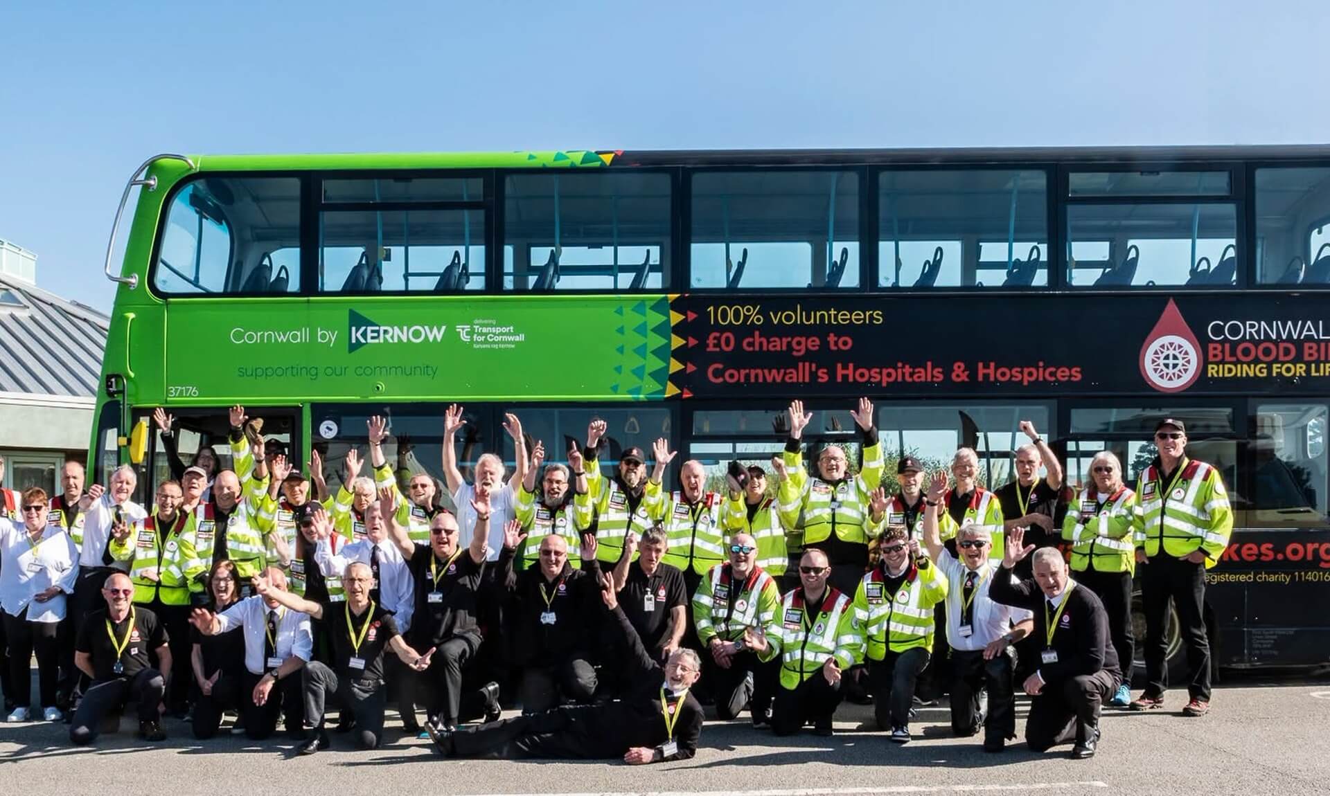 Cornwall Blood Bikes volunteers in hi-vis jackets cheering in front of a Kernow double decker bus in the charity's livery