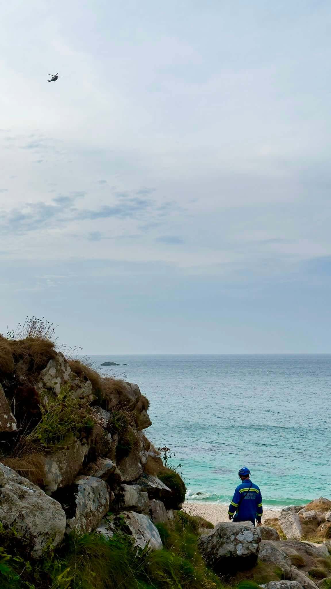 Coastguard team member watching a helicopter fly over the sea from a clifftop