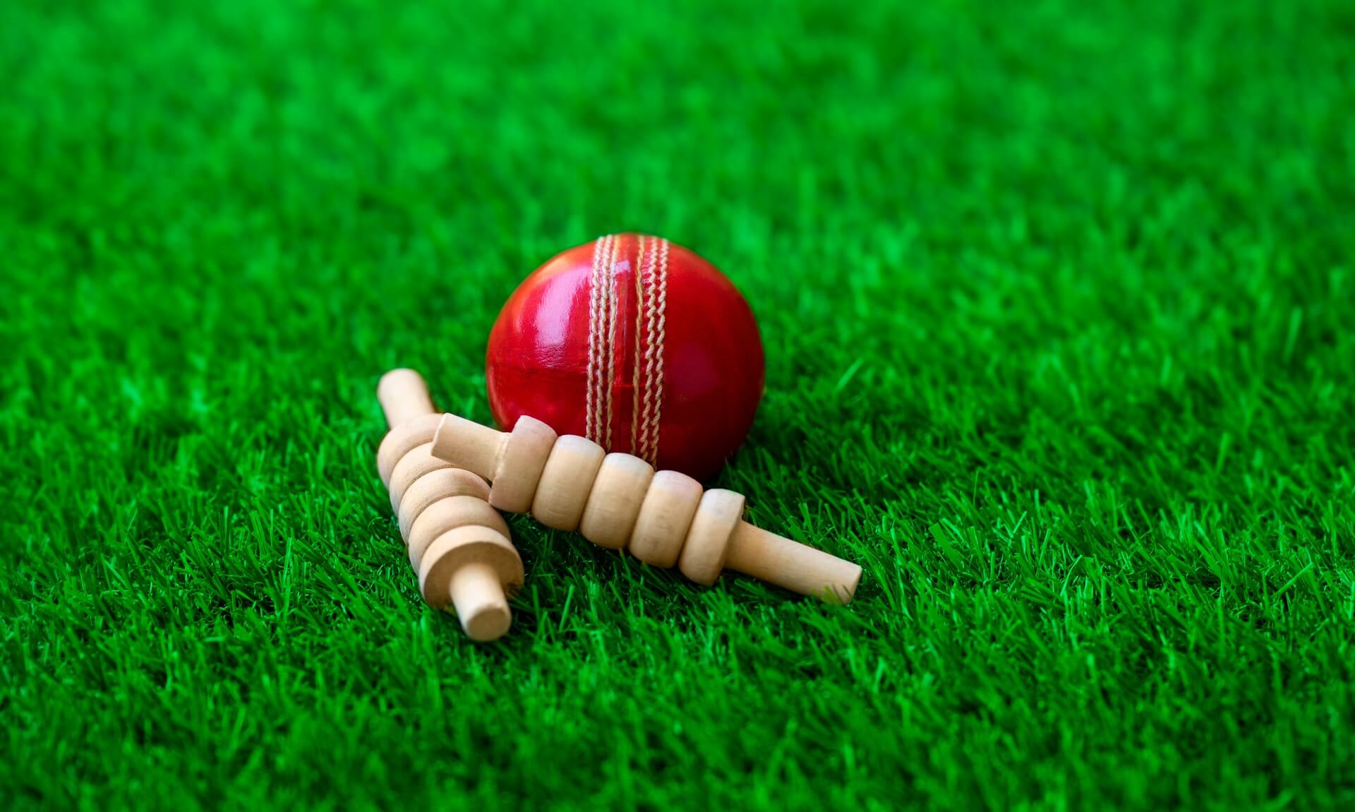 A red cricket ball resting on grass beside a set of knocked-over bails