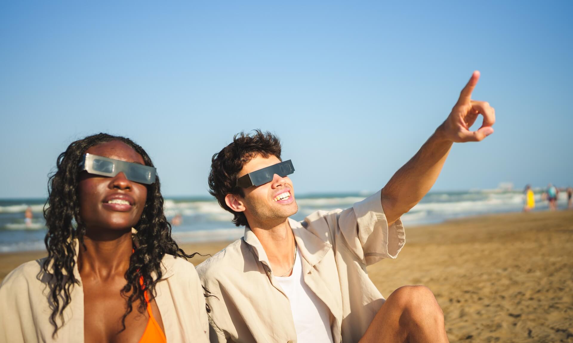 A couple on a beach wearing solar eclipse glasses, one pointing up at the sky
