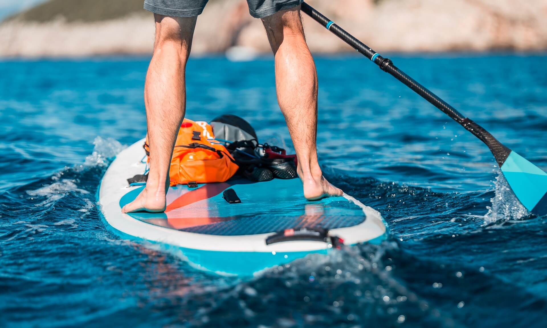 A paddleboarder standing on a stand-up paddleboard in blue open water off a rocky coast