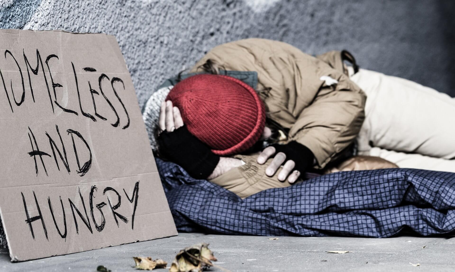 A cardboard sign reading Homeless and Hungry next to a person sleeping rough under blankets