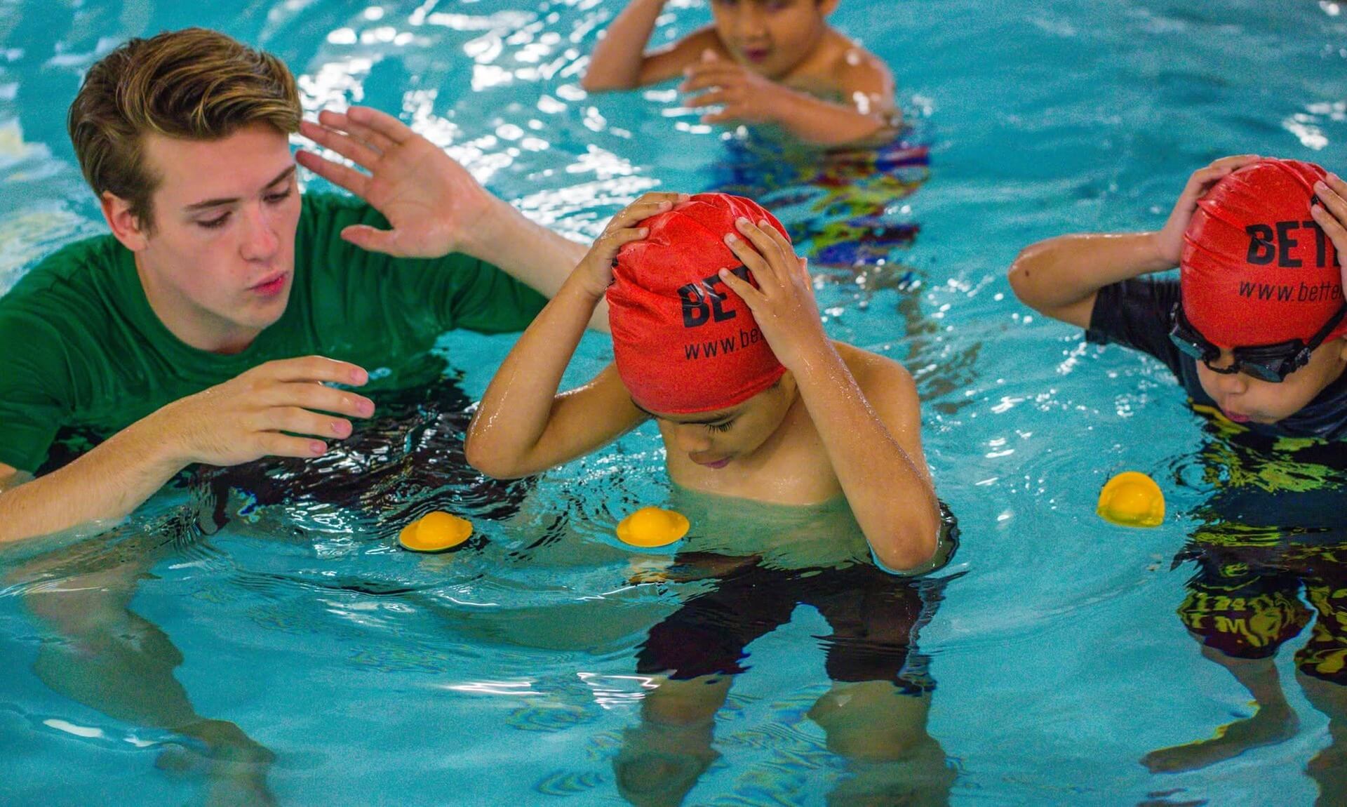 A swimming teacher instructing young children wearing red Better swim caps in a leisure centre pool