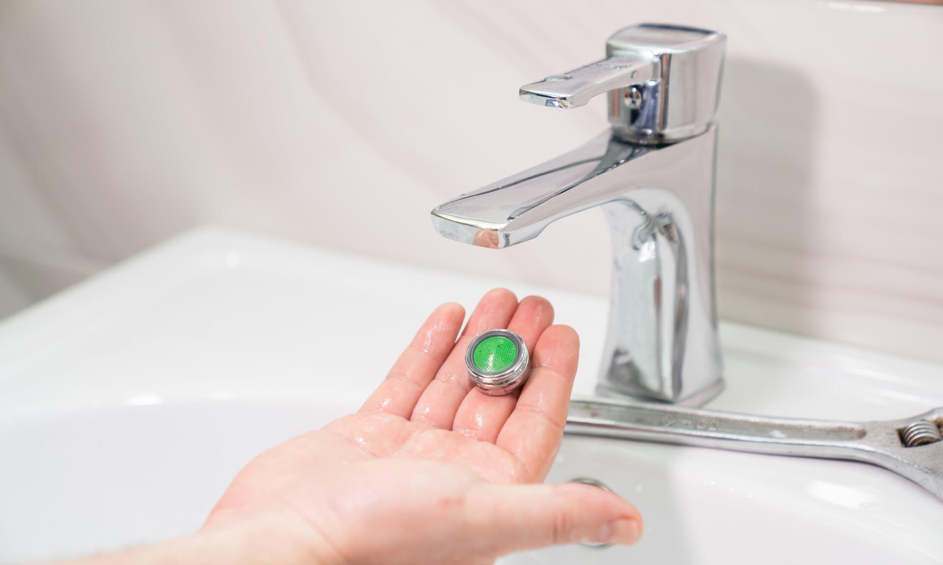 A hand holding a green water-saving tap aerator next to a chrome bathroom tap