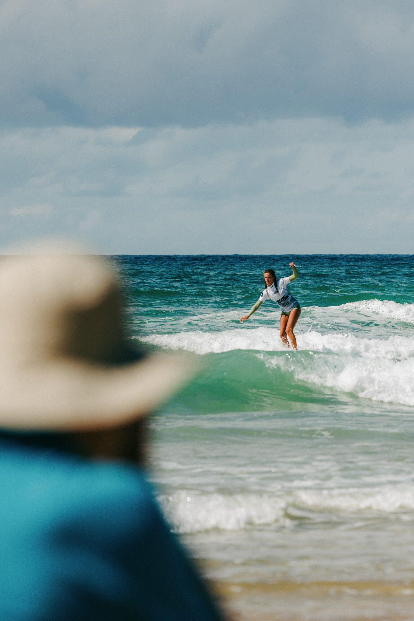 A spectator in a straw hat watches a Boardmasters Open competitor ride a wave at Fistral