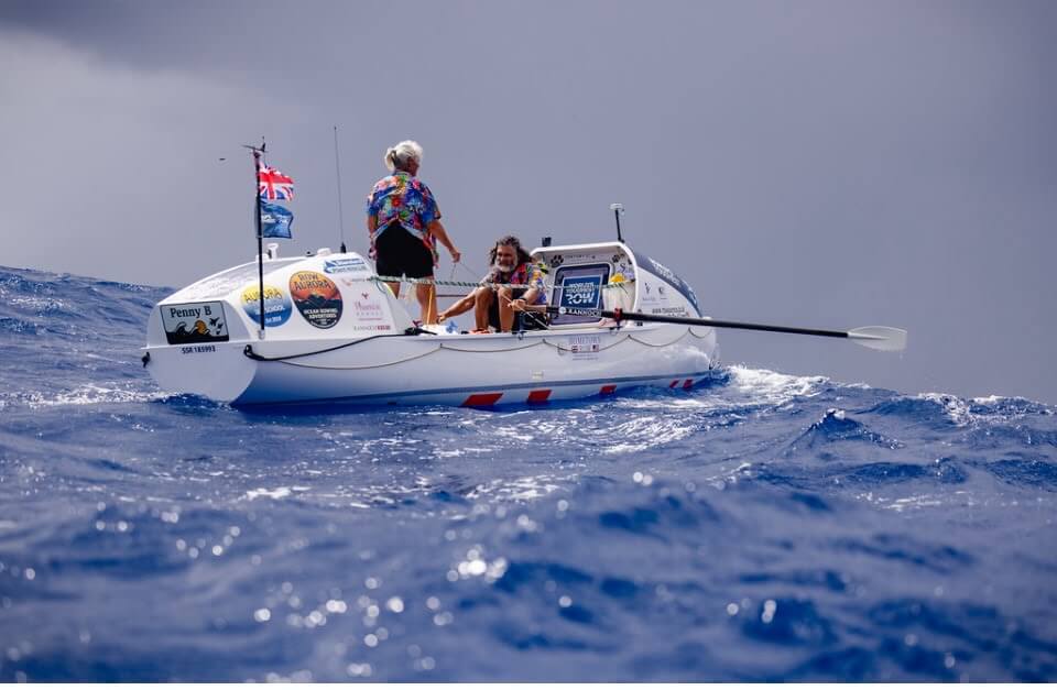 The ocean rowing boat Penny B under way at sea with one rower standing and one at the oars