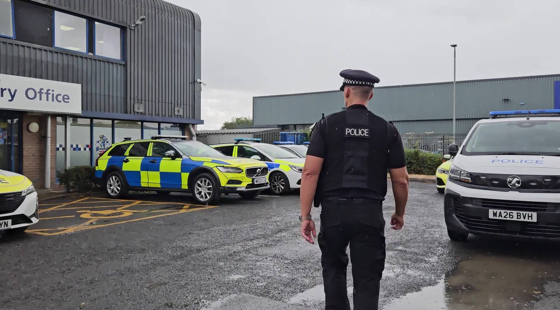 A police officer walking through a station car park lined with marked Devon and Cornwall Police vehicles