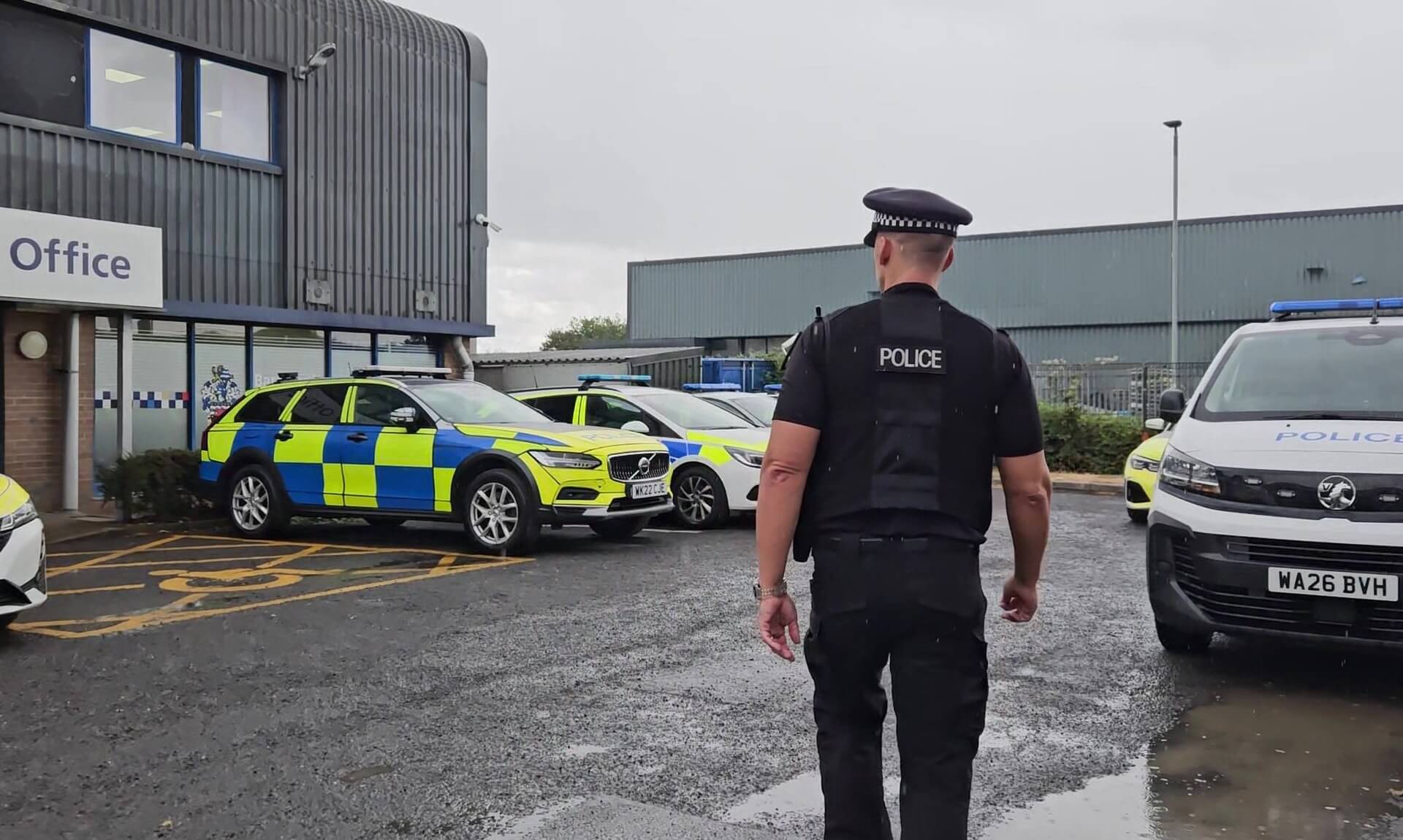 A police officer walking through a station car park lined with marked Devon and Cornwall Police vehicles