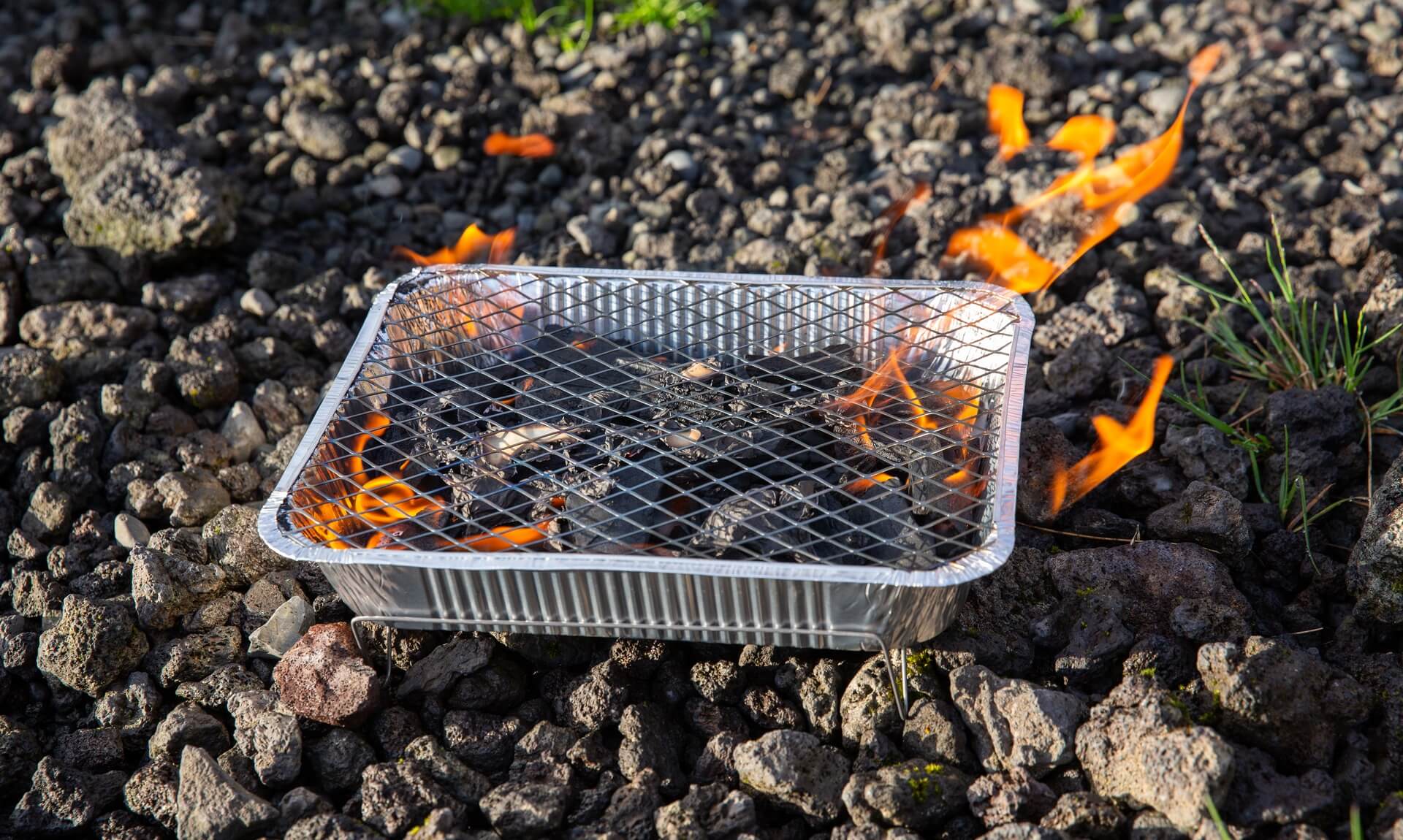 A disposable foil barbecue alight on stony ground with flames rising around it