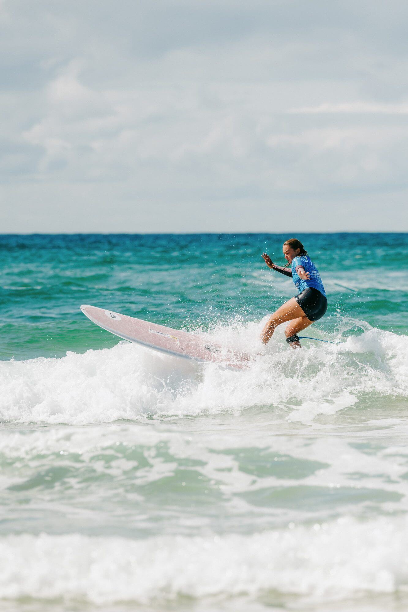 A competitor in a blue jersey turns hard on a pink longboard in the Fistral shorebreak