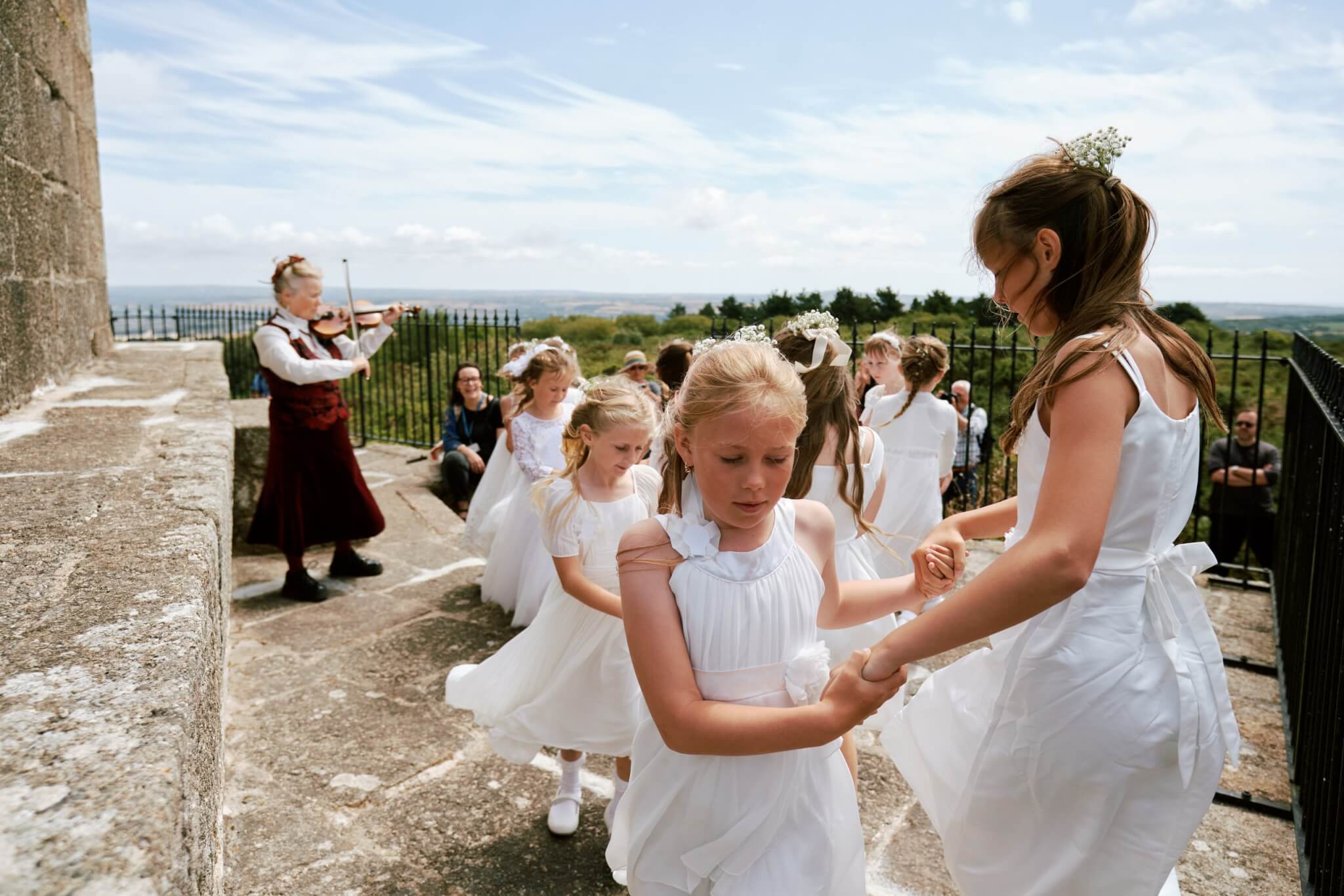 Maids in white dresses dance on the stone terrace at Knill's Monument with the fiddler and a sea view behind