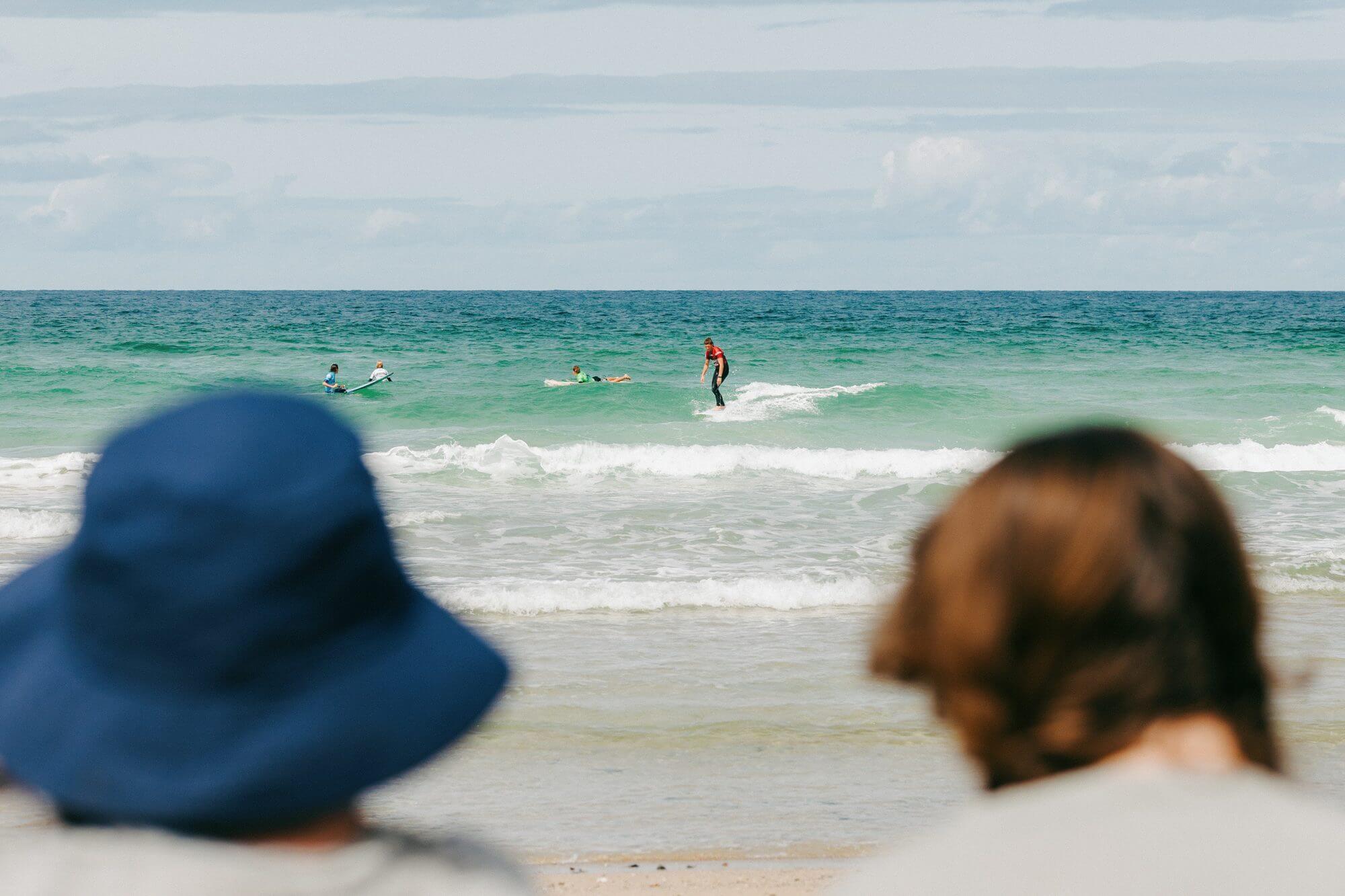Two spectators watch a surfer riding a wave at Fistral Beach