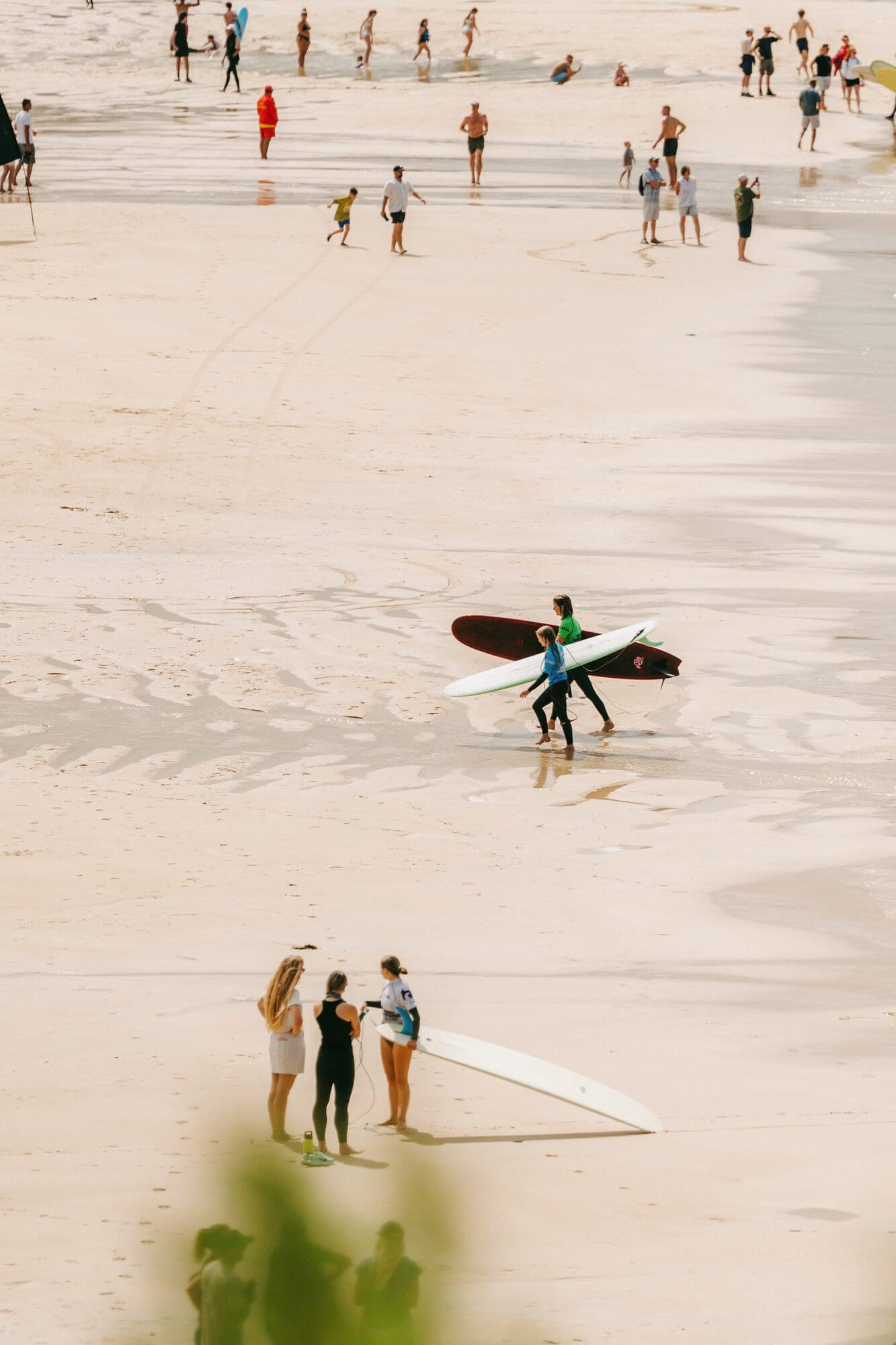Surfers carry their boards across Fistral Beach watched by spectators and coaches